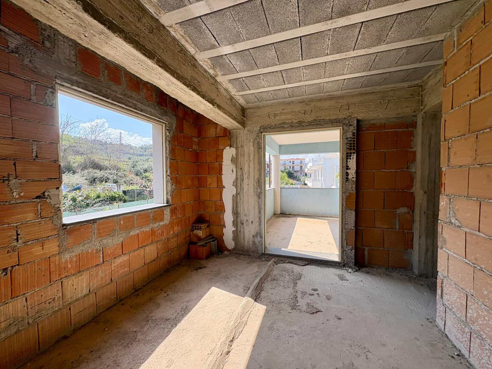 Vano interno di un appartamento in costruzione con pareti in mattoni rossi e finestre che offrono vista panoramica sulla natura circostante, pronto per essere rifinito e arredato.