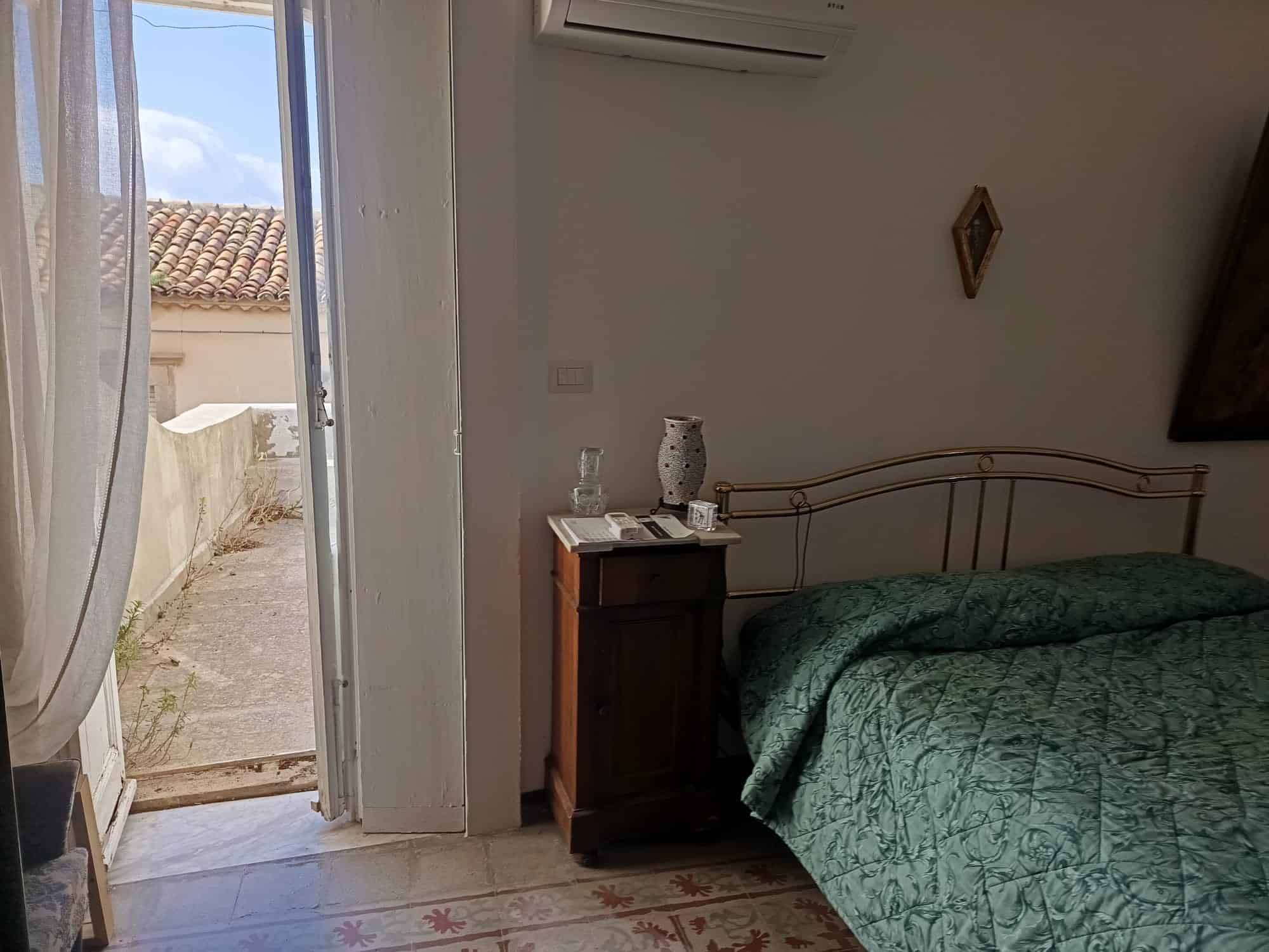 Riproduzione di una camera da letto luminosa con letto matrimoniale, comodino in legno, decorazioni semplici e accesso a un cortile esterno soleggiato, ideale per soggiorni rilassanti.