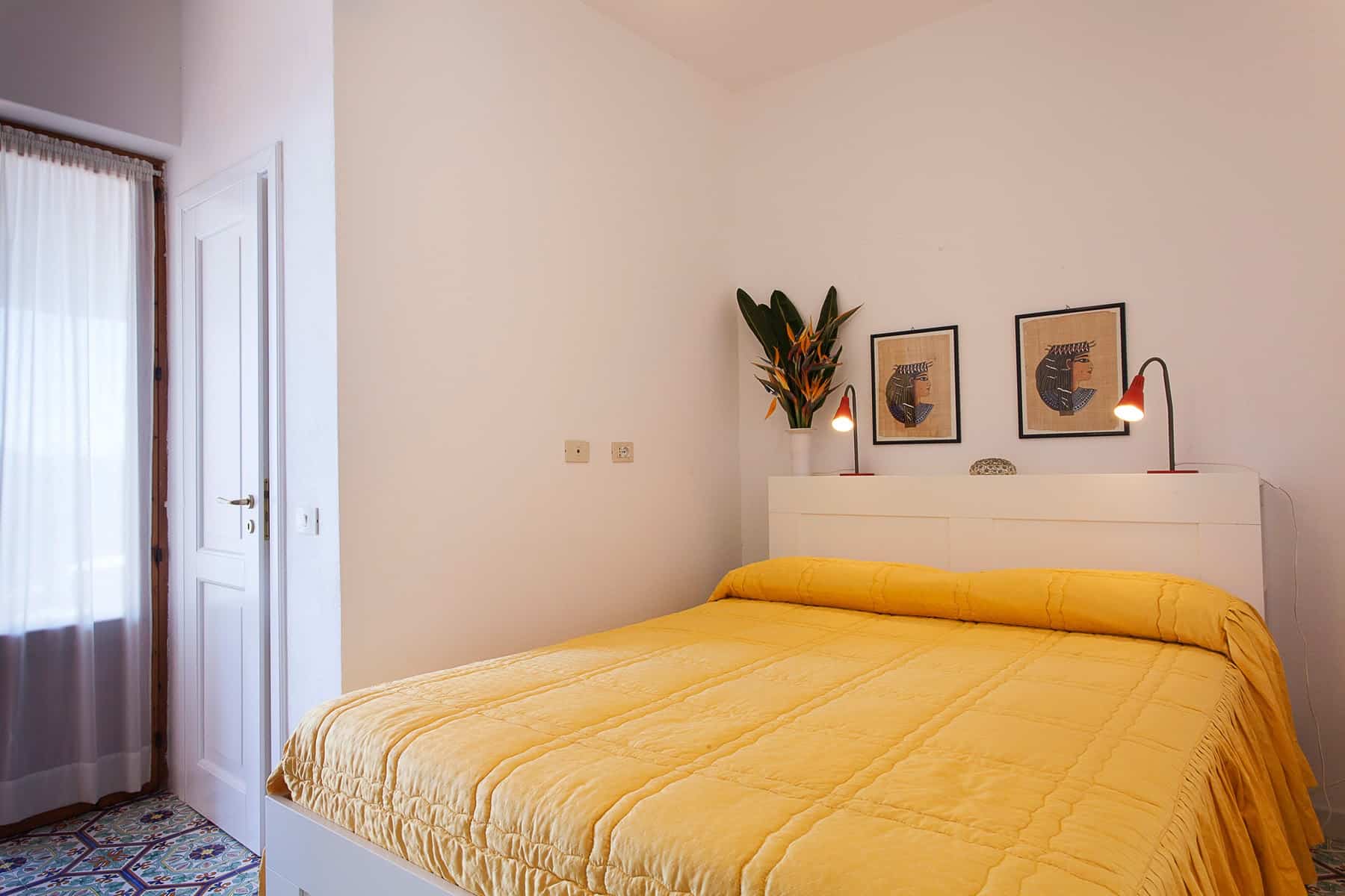 Ripetitore di una camera da letto moderna con pareti bianche, letto con coperta gialla, decorazioni artistiche e piante verdi, ambiente luminoso e accogliente.