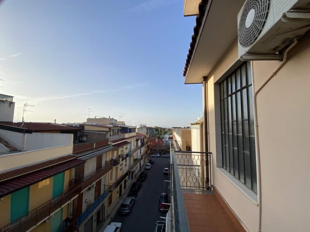 Balcone di un appartamento con vista su strada urbana, con edifici residenziali e auto parcheggiate, in una giornata soleggiata, in un quartiere tranquillo e ben servito.