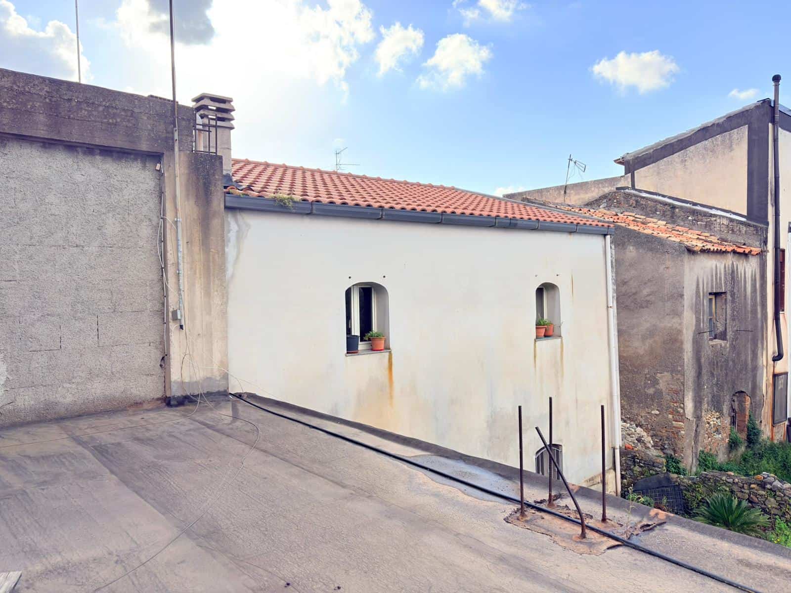 Il tetto di una casa con pendenza, con tegole rosse e una parete bianca, in un contesto urbano con edifici vicini e un cielo sereno.