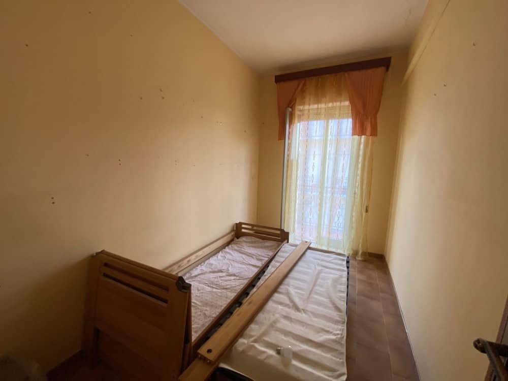 Letto singolo in camera con finestra luminosa, pareti beige e pavimento in legno, ideale per ambienti di soggiorno o camera da letto in affitto o vendita.