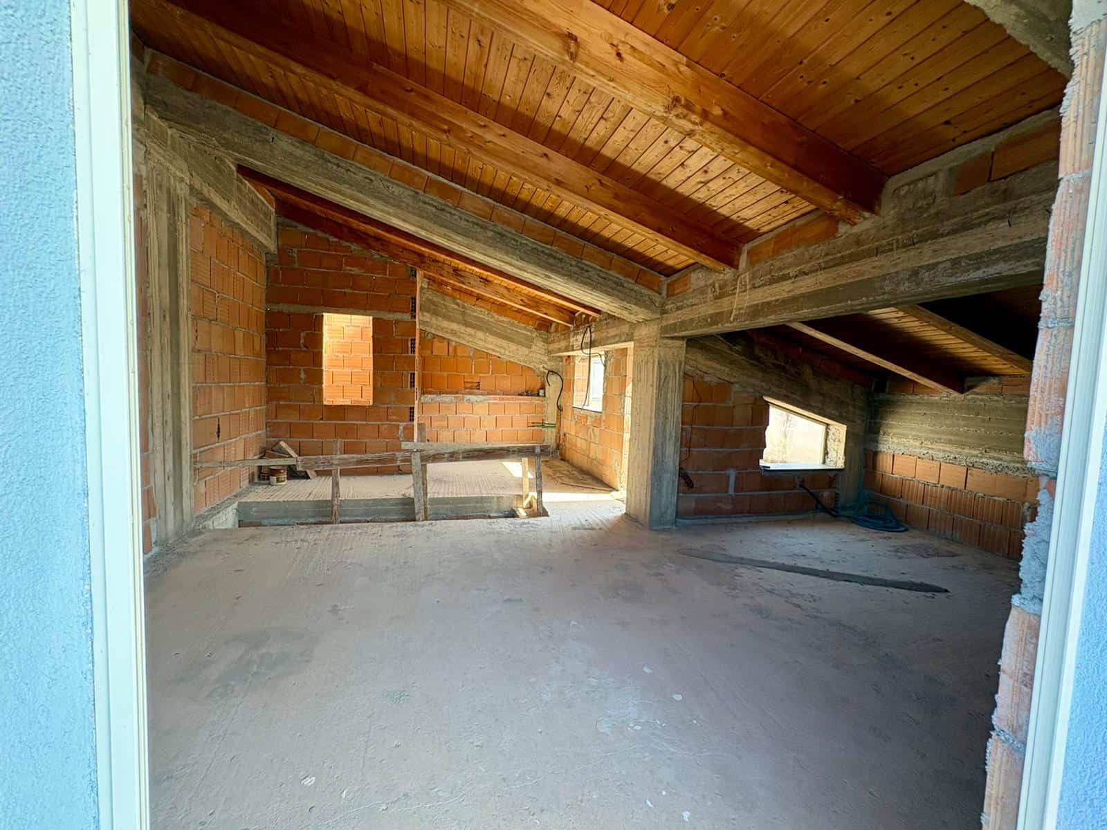 Ampia stanza in costruzione con pareti in mattoni e soffitto in legno, pronta per essere rifinita e personalizzata. Ideale come spazio abitativo o ufficio in fase di completamento.