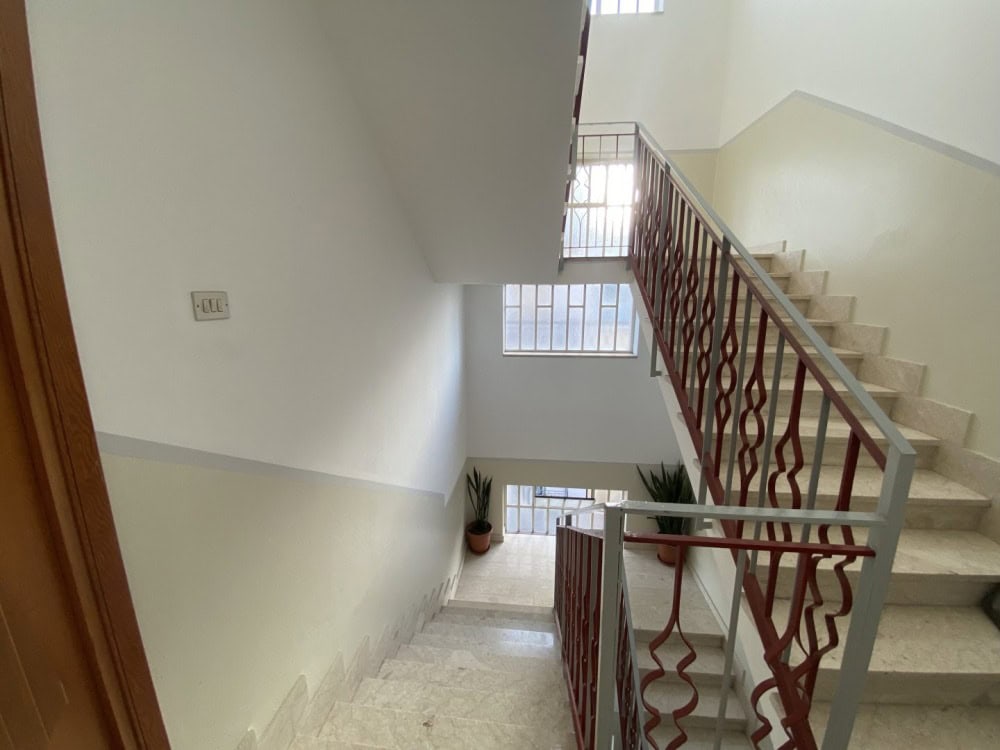 Escalier interno di un edificio residenziale con ringhiera in ferro battuto e pareti bianche, illuminato da luce naturale proveniente dalle finestre.