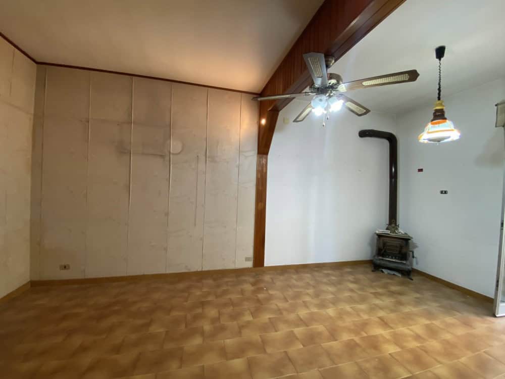 Interno di una stanza spaziosa con soffitto inclinato, ventilatore a soffitto, camino in ghisa e illuminazione sospesa, ideale per soggiorno o zona living.