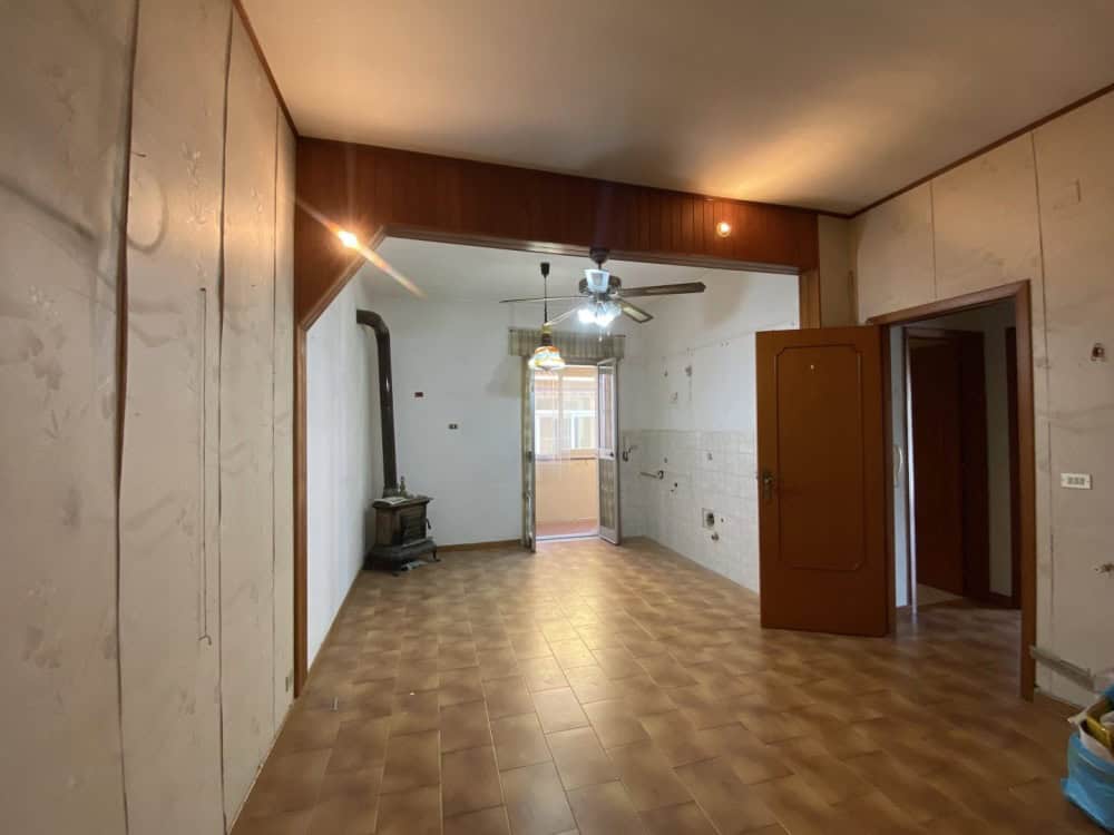 Ampia stanza vuota con pareti in cartongesso, pavimento in piastrelle e soffitto con ventilatore e luci, pronta per ristrutturazione o arredamento, ideale come soggiorno o spazio living.