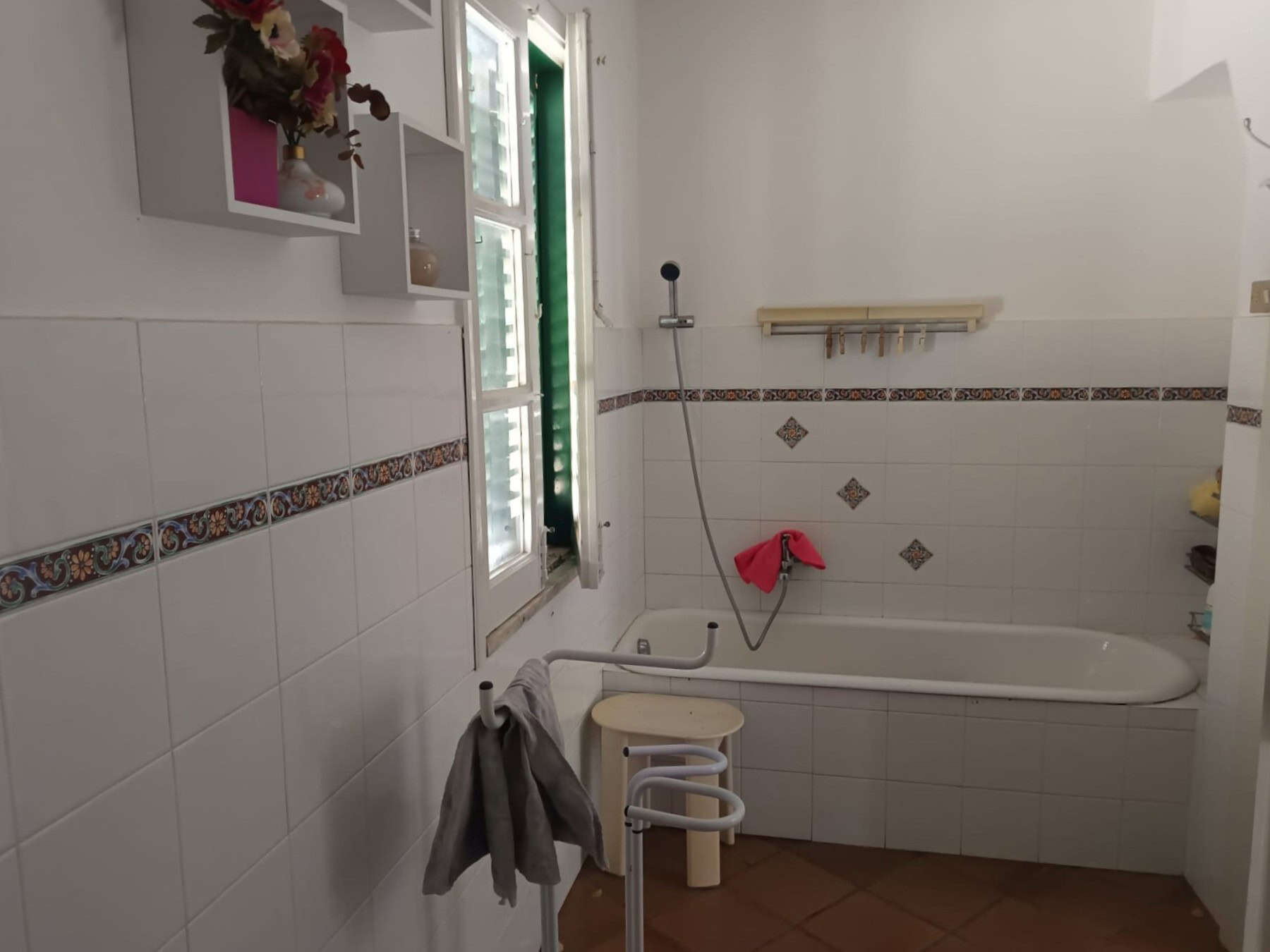 Ampia stanza da bagno con vasca, finestra con persiana, pareti decorate con piastrelle bianche e dettagli colorati, accessori come asciugamani e mensole per prodotti da bagno.