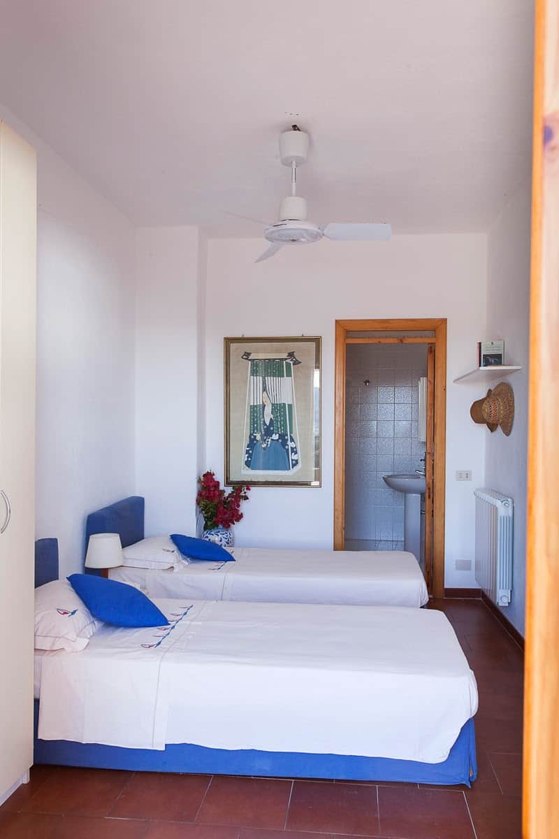 Letto doppio con lenzuola bianche e cuscini blu, stanza luminosa con pareti bianche, decorazioni marine e bagno visibile in background, ideale per soggiorni in località di mare.