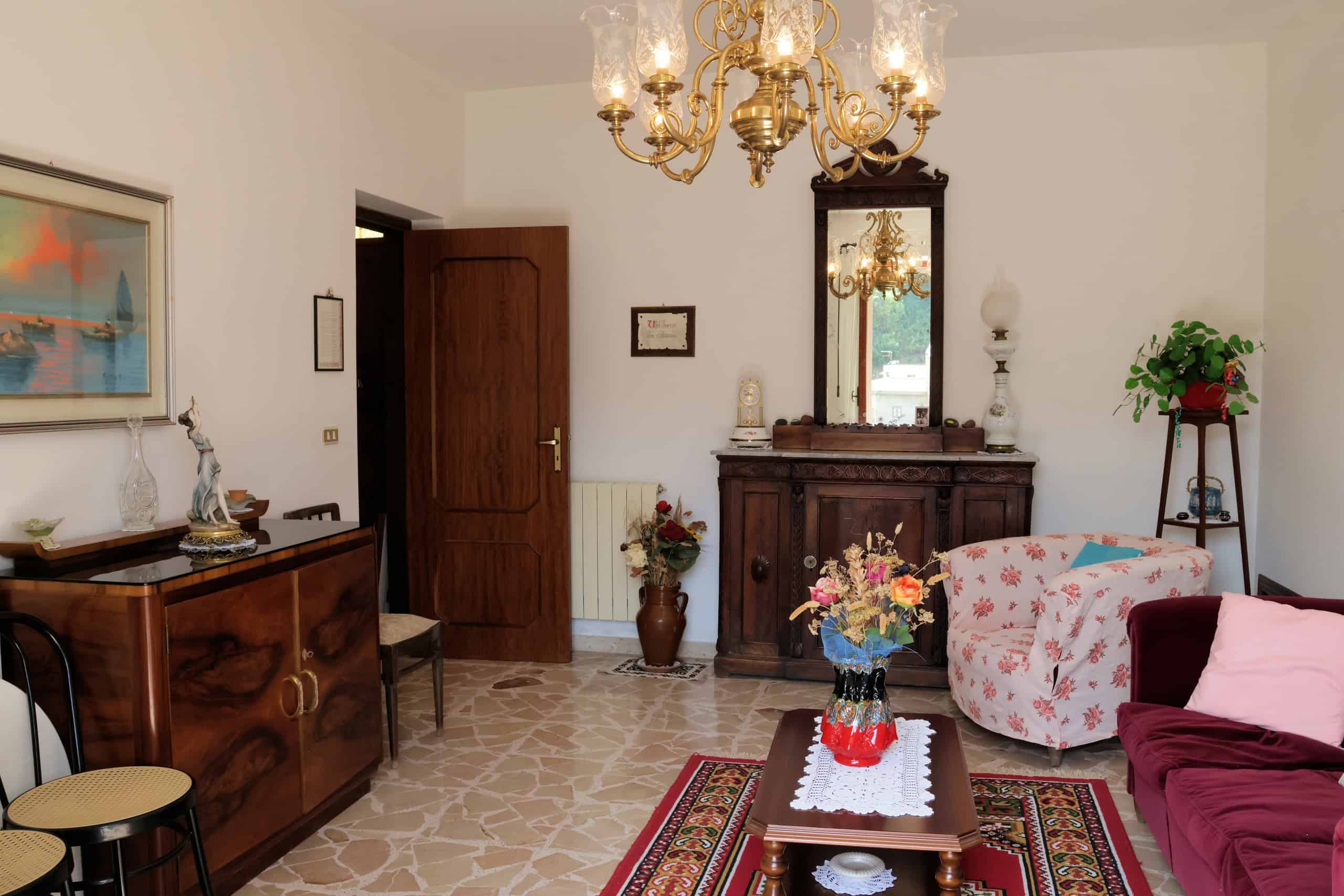 Elegante soggiorno con mobili in legno, divano con cuscini, lampadario classico e decorazioni floreali, ideale per vivere momenti di relax in un ambiente accogliente e raffinato.