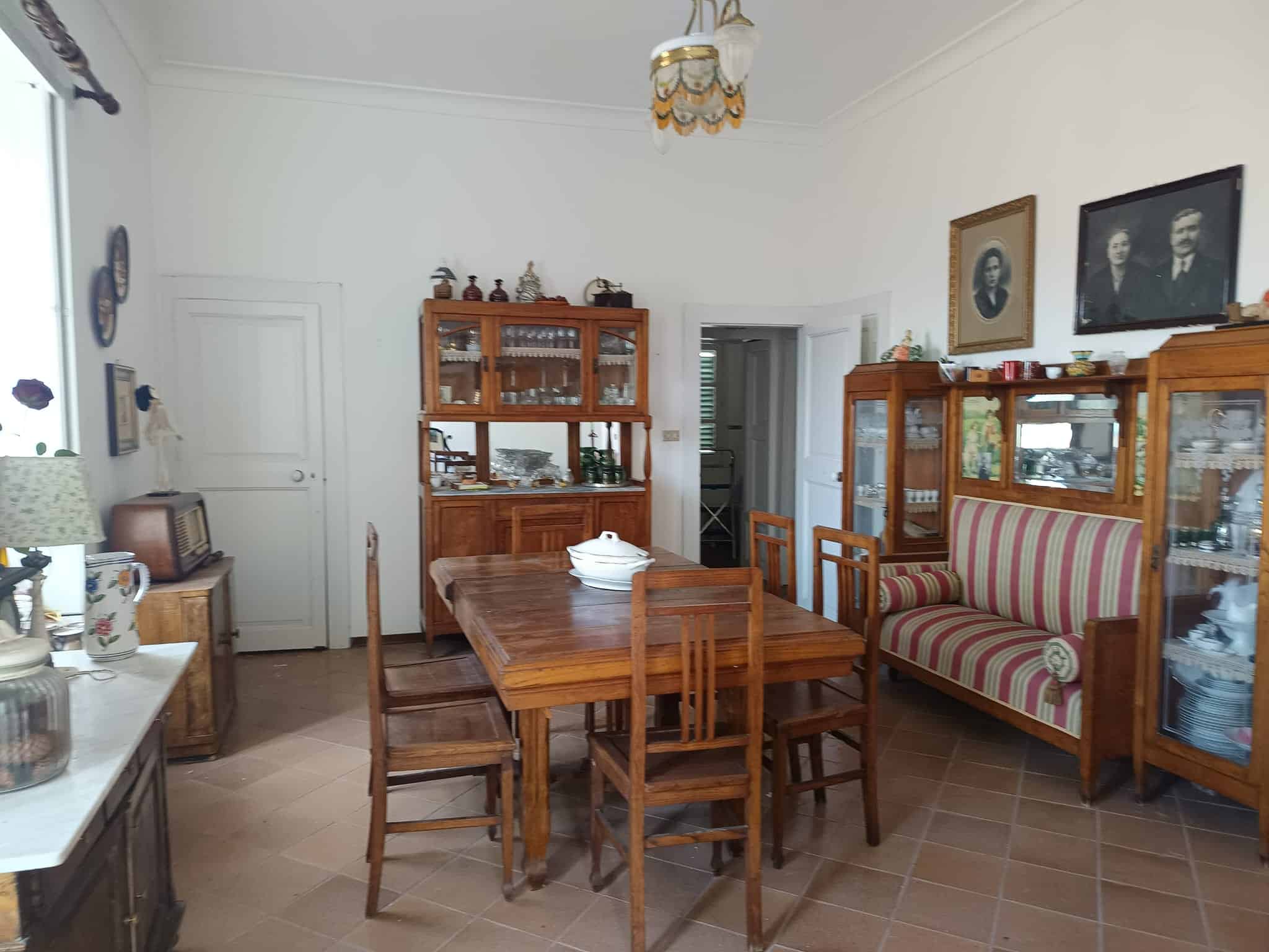 Interno di una sala da pranzo con mobili in legno, tavolo centrale e sedie, decorazioni vintage e fotografie alle pareti, atmosfera accogliente e classica.