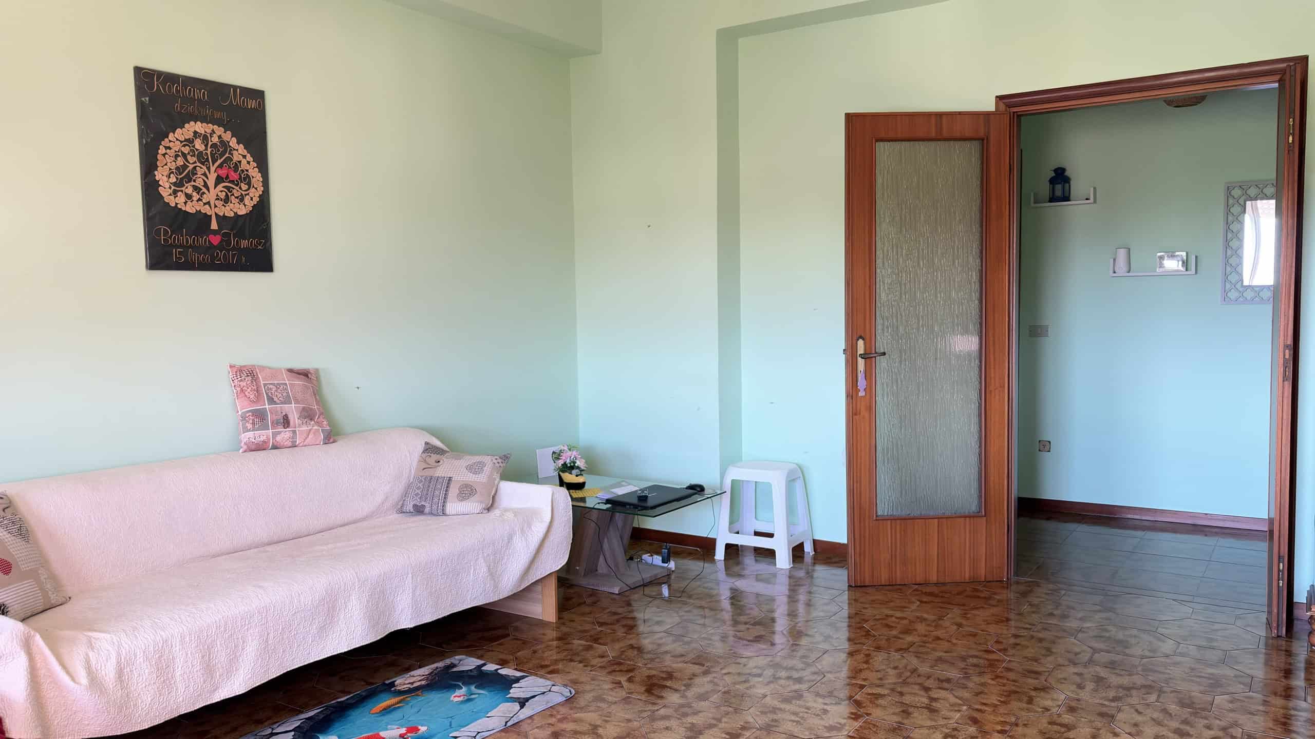 Ampia stanza soggiorno con divano beige, pareti verde pastello e pavimento in parquet, porta in legno con vetro decorato, decorazioni eleganti e ambiente luminoso.