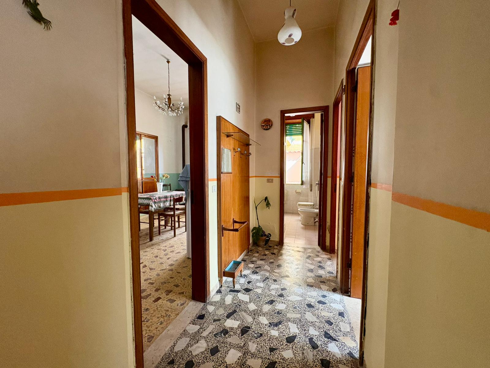Corridoio interno di una casa con pavimento in mosaico, pareti beige e porte in legno, illuminato da luce naturale e lampadario centrale.