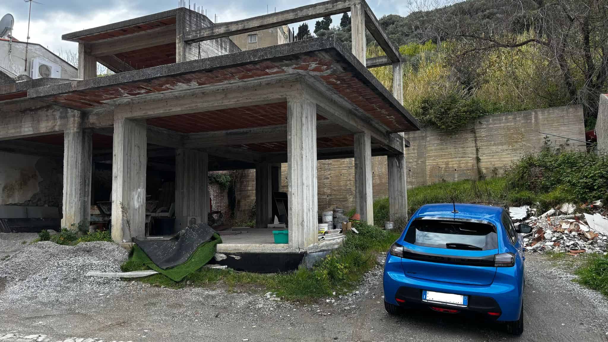 Auto sportivo blu parcheggiato davanti a una struttura in costruzione con pilastri di cemento e tetto in legno, in un'area in pendenza circondata da vegetazione e muri di contenimento, rappresentando.
