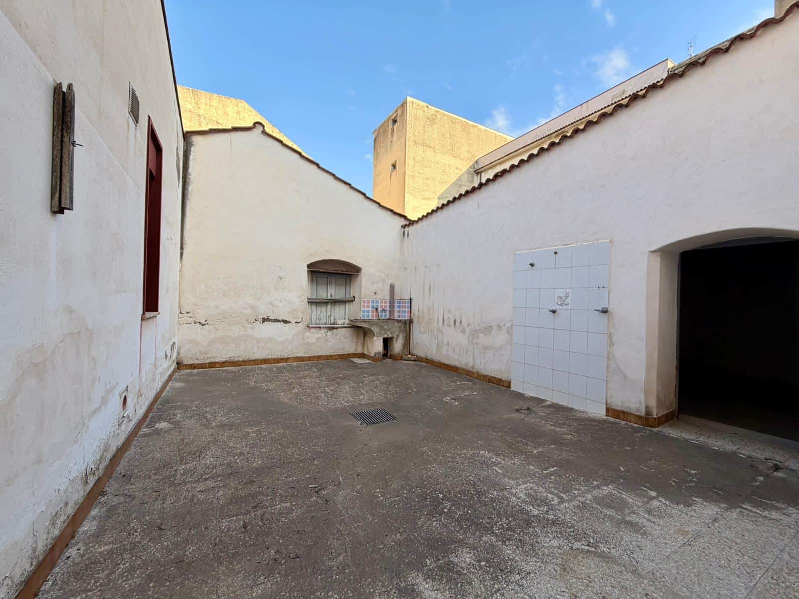 Cortile esterno di una proprietà immobiliare con pareti bianche, pavimento in cemento e un'area coperta, ideale per attività all'aperto o parcheggio.