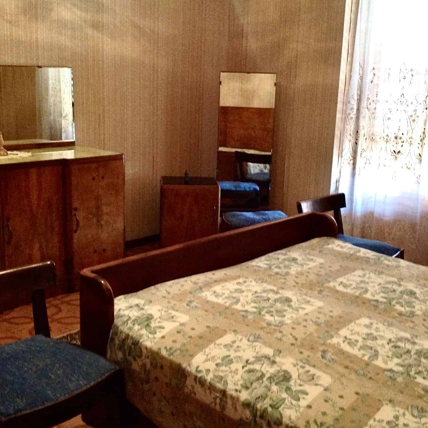 Una stanza da letto con mobili in legno, un letto matrimoniale con copriletto floreale, specchio e sedie, illuminata dalla luce naturale attraverso una finestra con tende leggere.