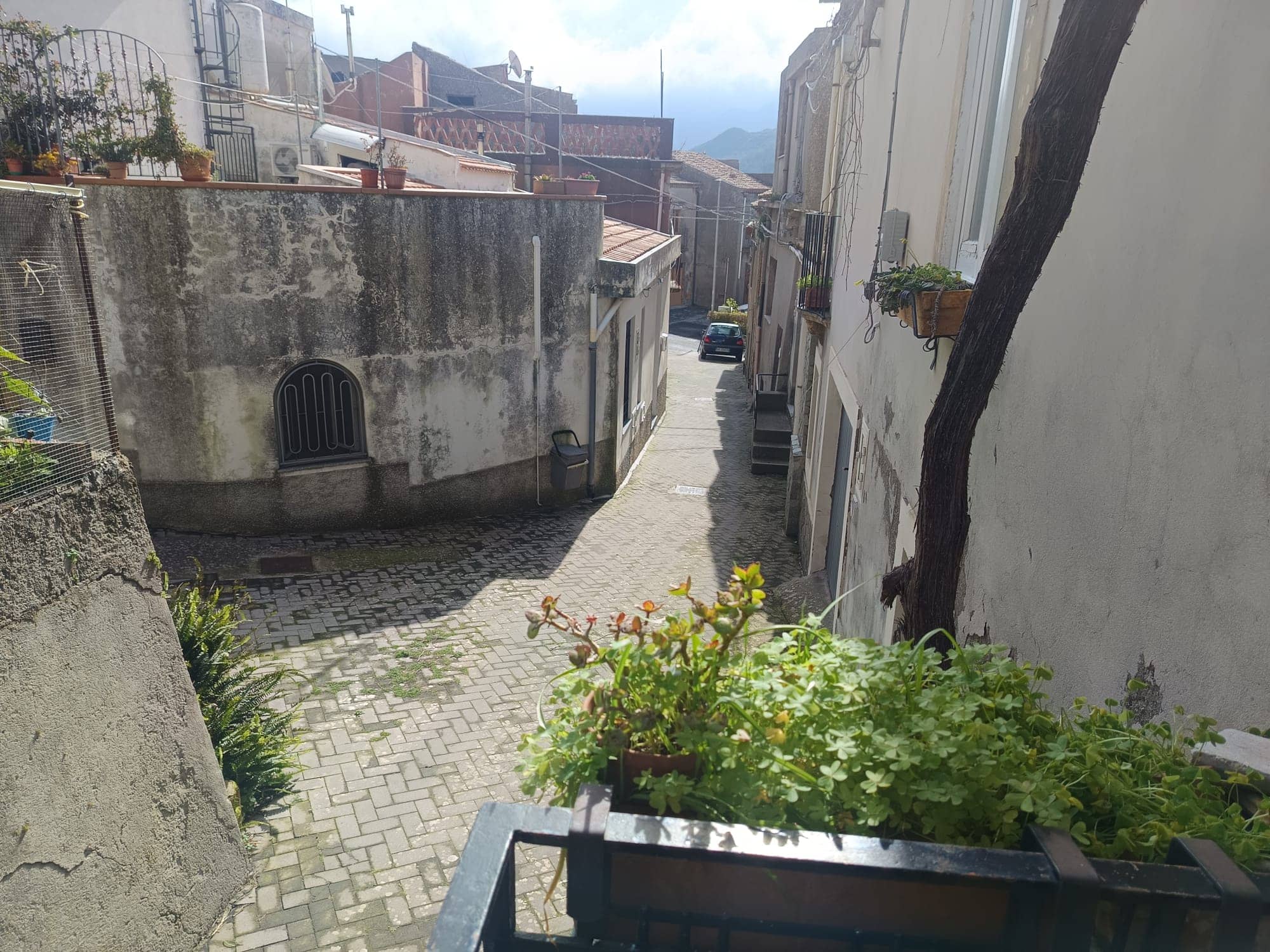 Barriera di strada stretta con edifici storici e piante verdi, vista verso un vicolo di città con case antiche e montagne sullo sfondo, atmosfera tranquilla e pittoresca.