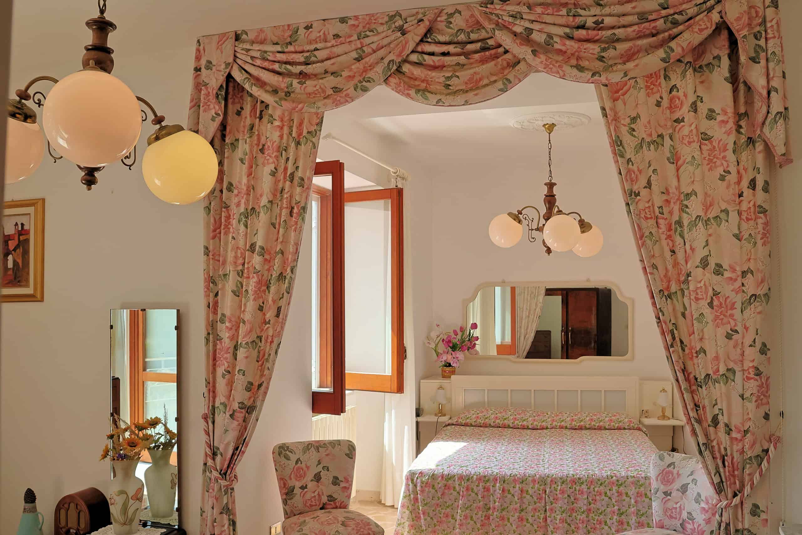 Una stanza da letto accogliente con tende floreali, letto matrimoniale e decorazioni vintage, ideale per creare un ambiente caldo e invitante in stile classico. Perfetta per case di charme e ambienti.