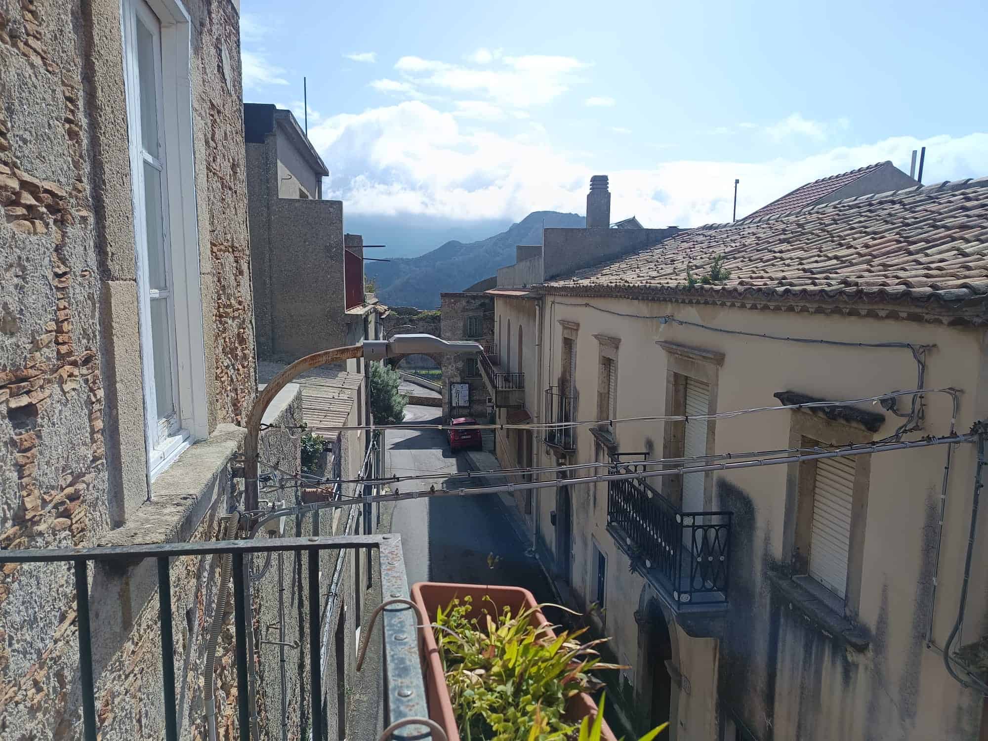 Balcone con vista sulle case storiche e montagne sullo sfondo, in un paesaggio urbano italiano, con linee elettriche e dettagli architettonici tradizionali.