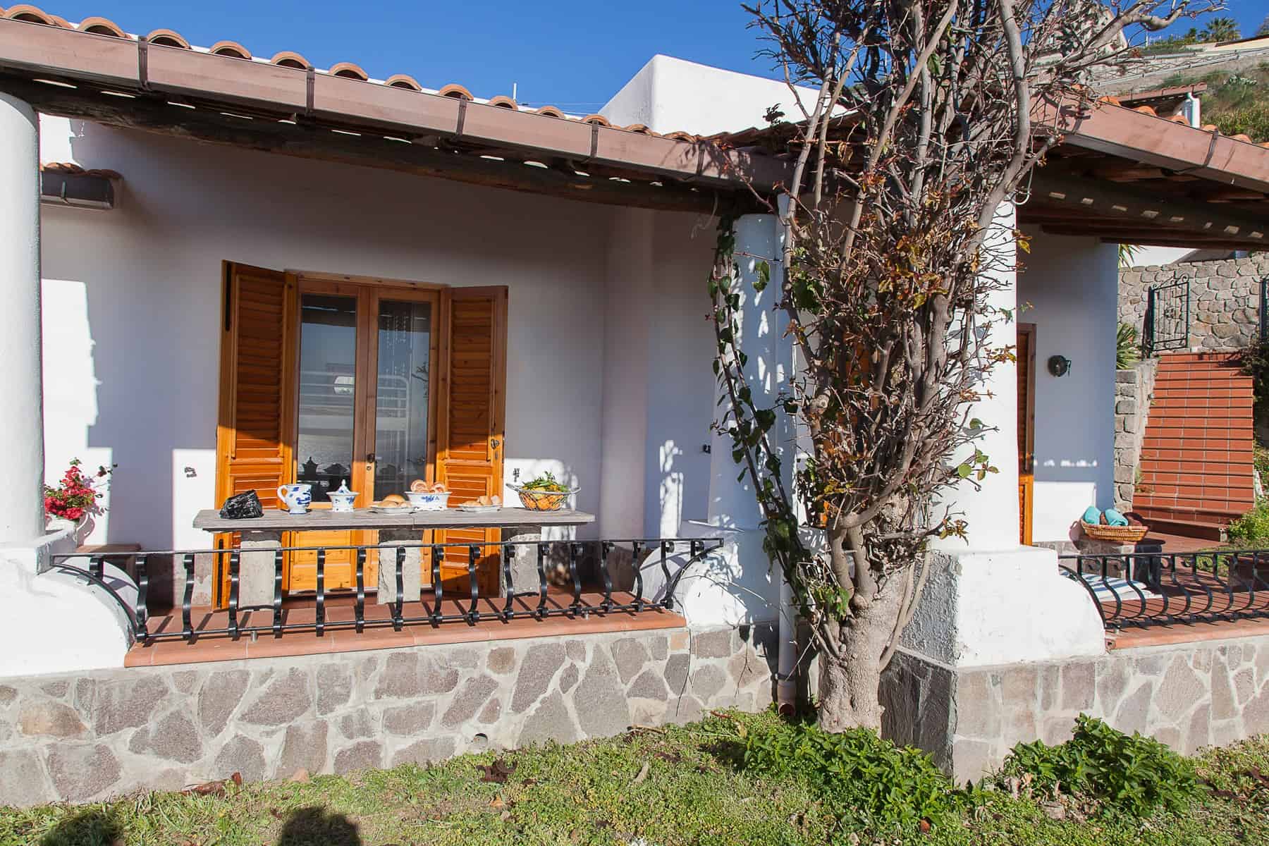 Finestra con persiane in legno e terrazza esterna di una casa tradizionale in stile mediterraneo, con decorazioni floreali e arredi esterni, immersa in un ambiente tranquillo e soleggiato.