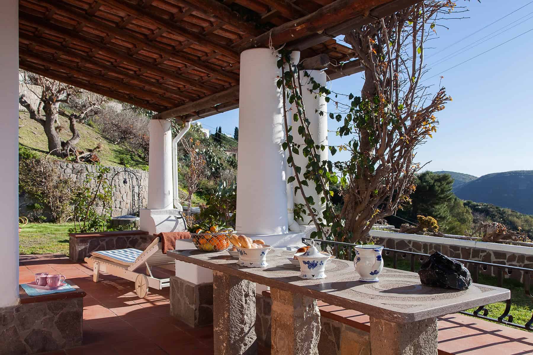 Veranda con vista panoramica, tavolo in pietra con oggetti decorativi, sedie e piante, ambiente esterno tranquillo e accogliente in campagna.