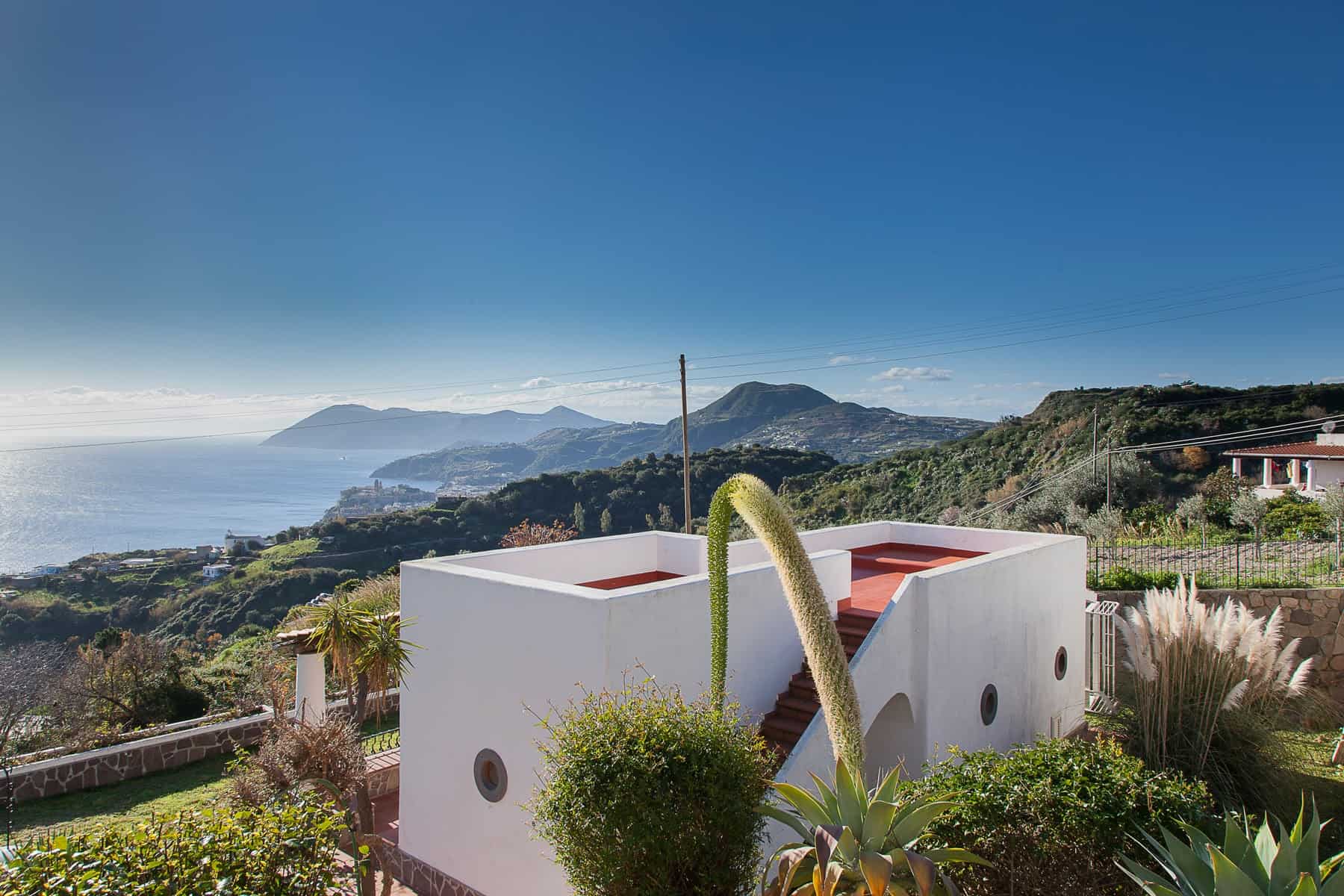 Casa moderna bianca con terrazza panoramica, vista mare e montagne, giardino con piante tropicali, in zona collinare soleggiata, stile minimalista, ideale per vacanze relax in Italia.