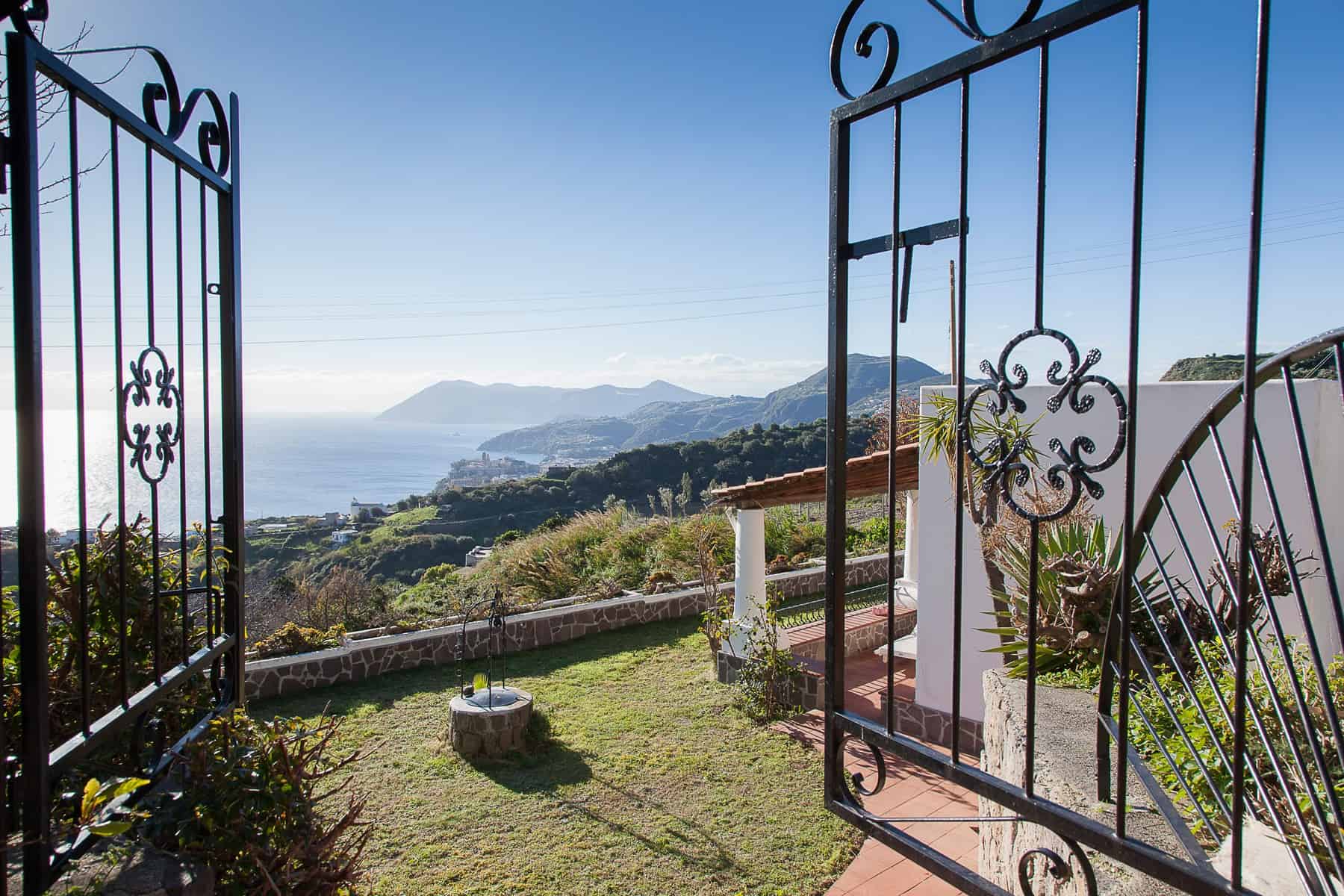 Cancello in ferro battuto che si apre su un giardino con vista panoramica sul mare e sulle colline circostanti, in un ambiente tranquillo e suggestivo.