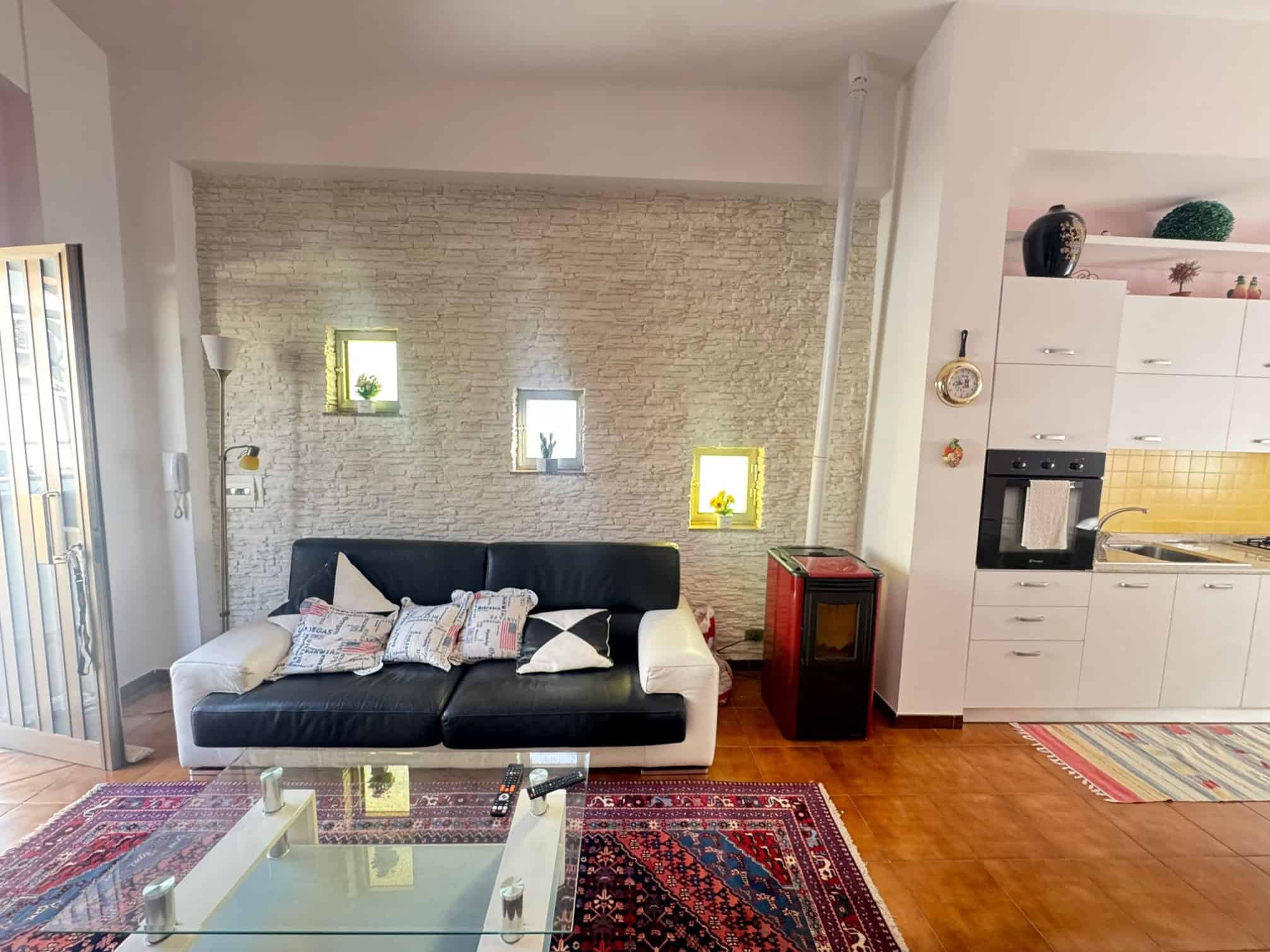 Sedia moderna in soggiorno con parete in pietra, decorazioni colorate e area cucina aperta con elettrodomestici integrati, ambiente accogliente e luminoso.