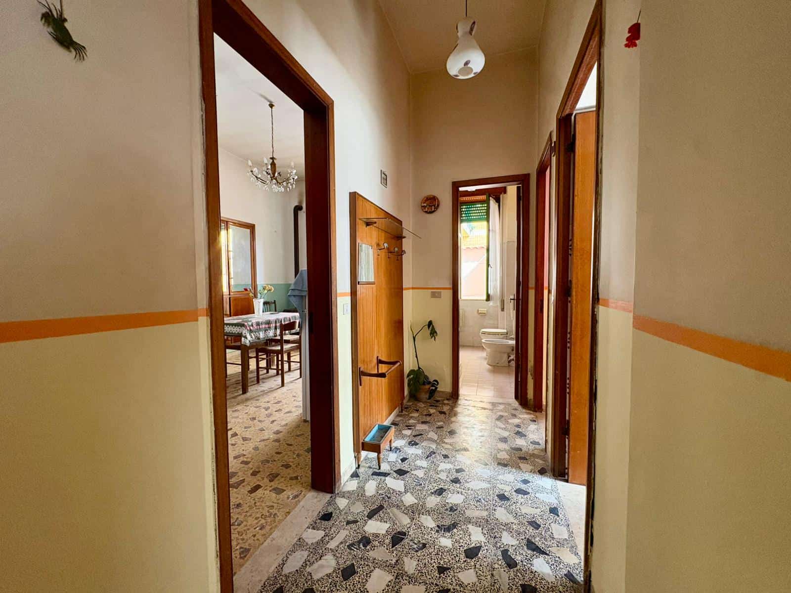 Ampio corridoio interno di una casa con pavimento in mosaico, pareti beige con una striscia arancione, porte in legno e finestra che permette l'ingresso di luce naturale.