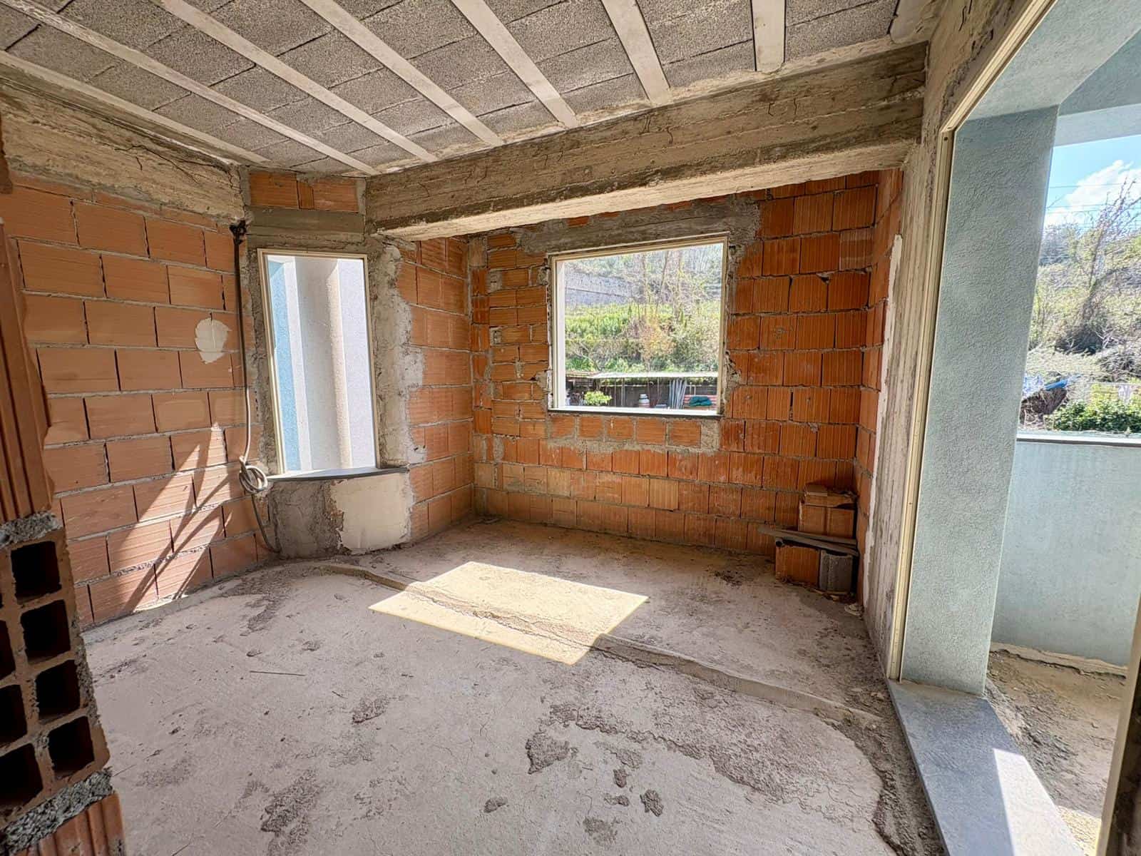 Vano interno di una casa in costruzione con pareti in mattoni e finestre, pronto per essere rifinito e arredato, con vista sul paesaggio naturale circostante.