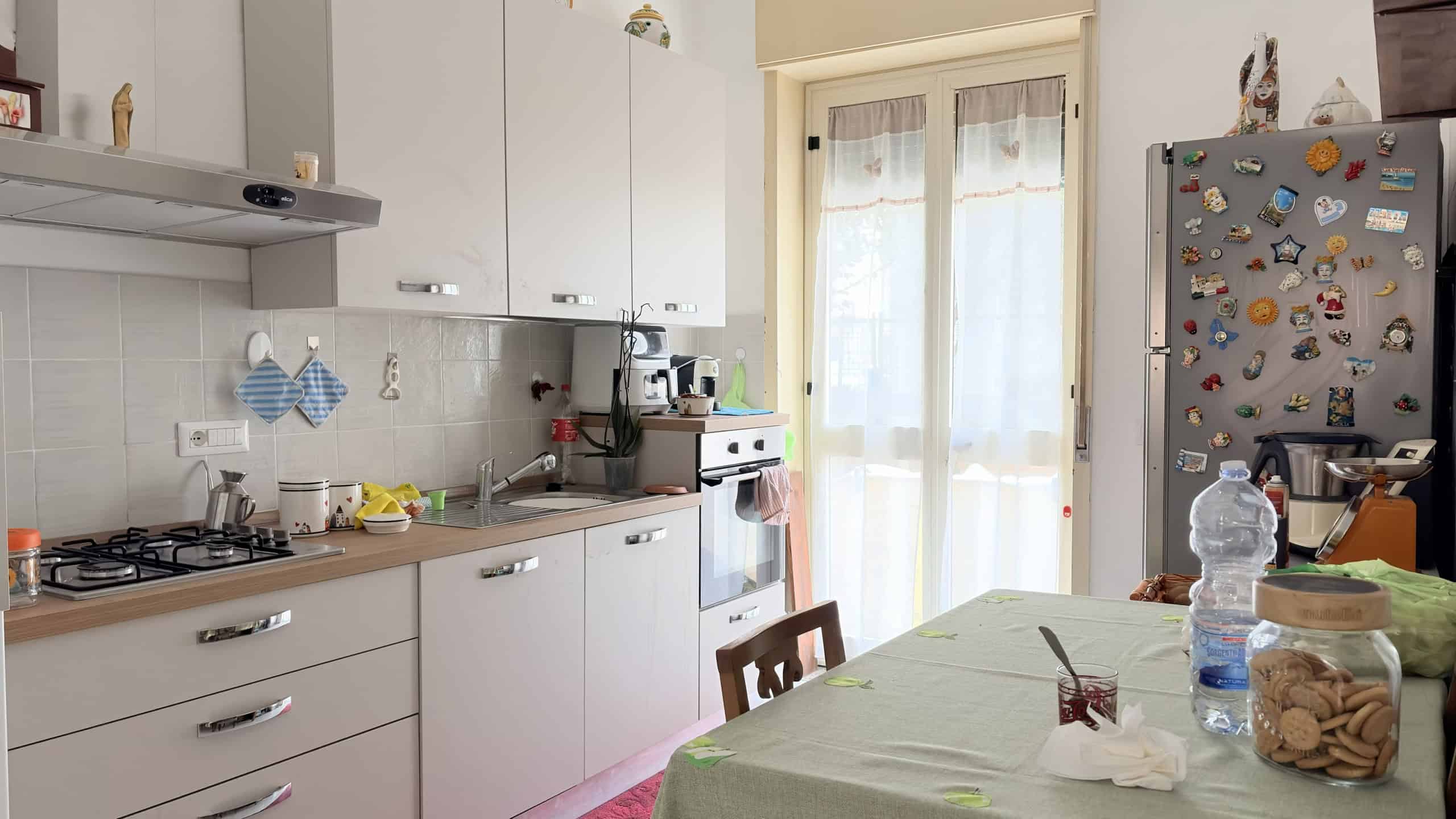 Pannello di cucina moderna con elettrodomestici, tavolo con alimenti e accessori, illuminata da luce naturale attraverso una porta finestra.