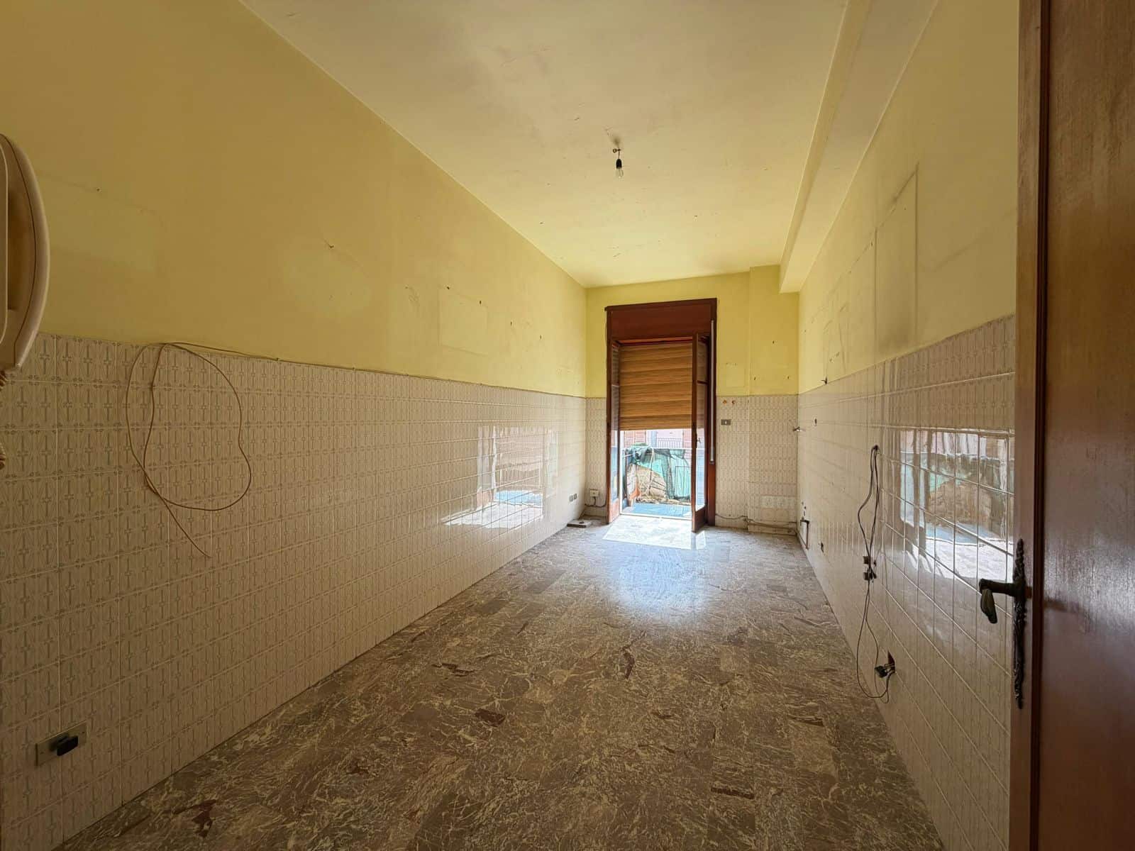 Ampia stanza da ristrutturare con pareti gialle e rivestimenti in piastrelle bianche, luce naturale proveniente dalla porta finestra, pavimento in marmo, ideale per cucina o soggiorno.