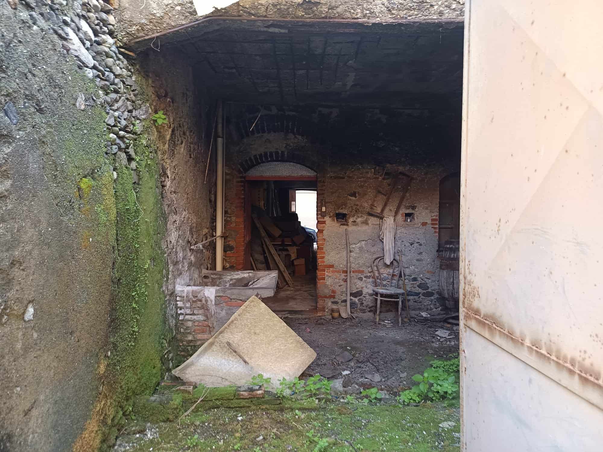 Abbandono di un edificio storico con pareti in pietra e mattoni, ingresso aperto e ambiente interno trascurato, con detriti e vegetazione che crescono sul pavimento.