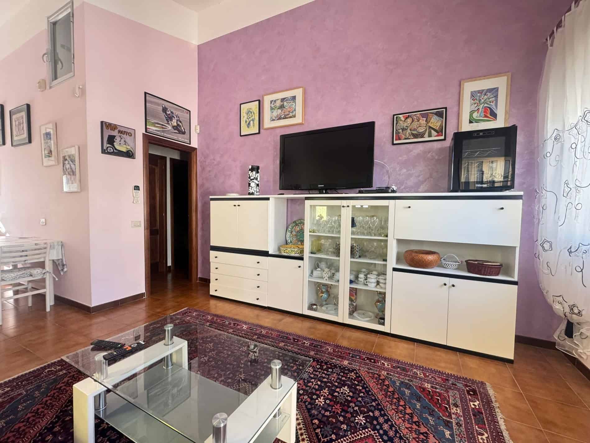 Interno soggiorno con mobile TV bianco, pareti rosa e decorazioni artistiche, pavimento in parquet e tappeto persiano, ambiente accogliente e luminoso.