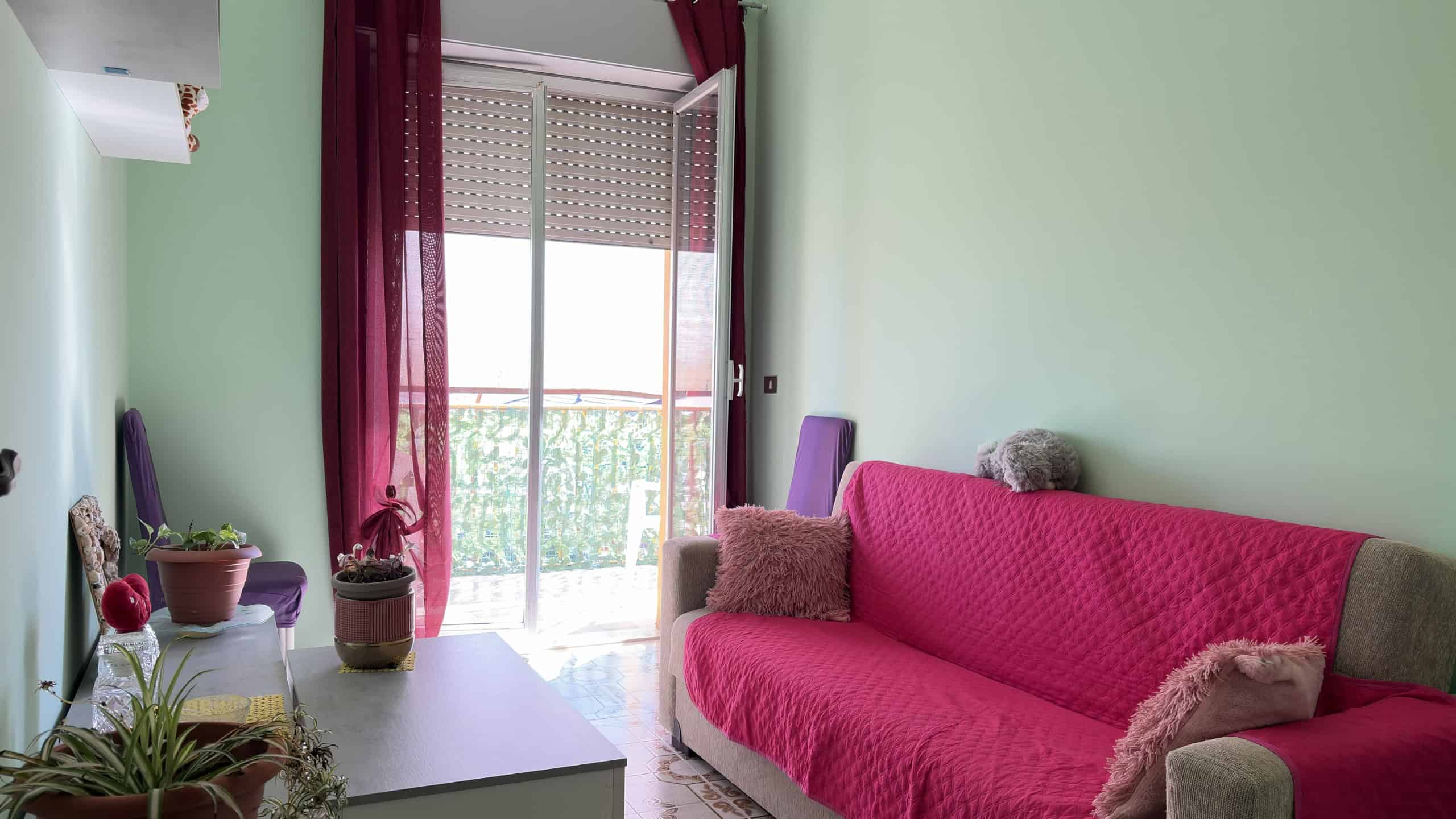 Sedia viola e divano rosa in soggiorno luminoso con balcone e tende trasparenti, decorazioni floreali e piante in vaso, ambiente accogliente e ben illuminato.
