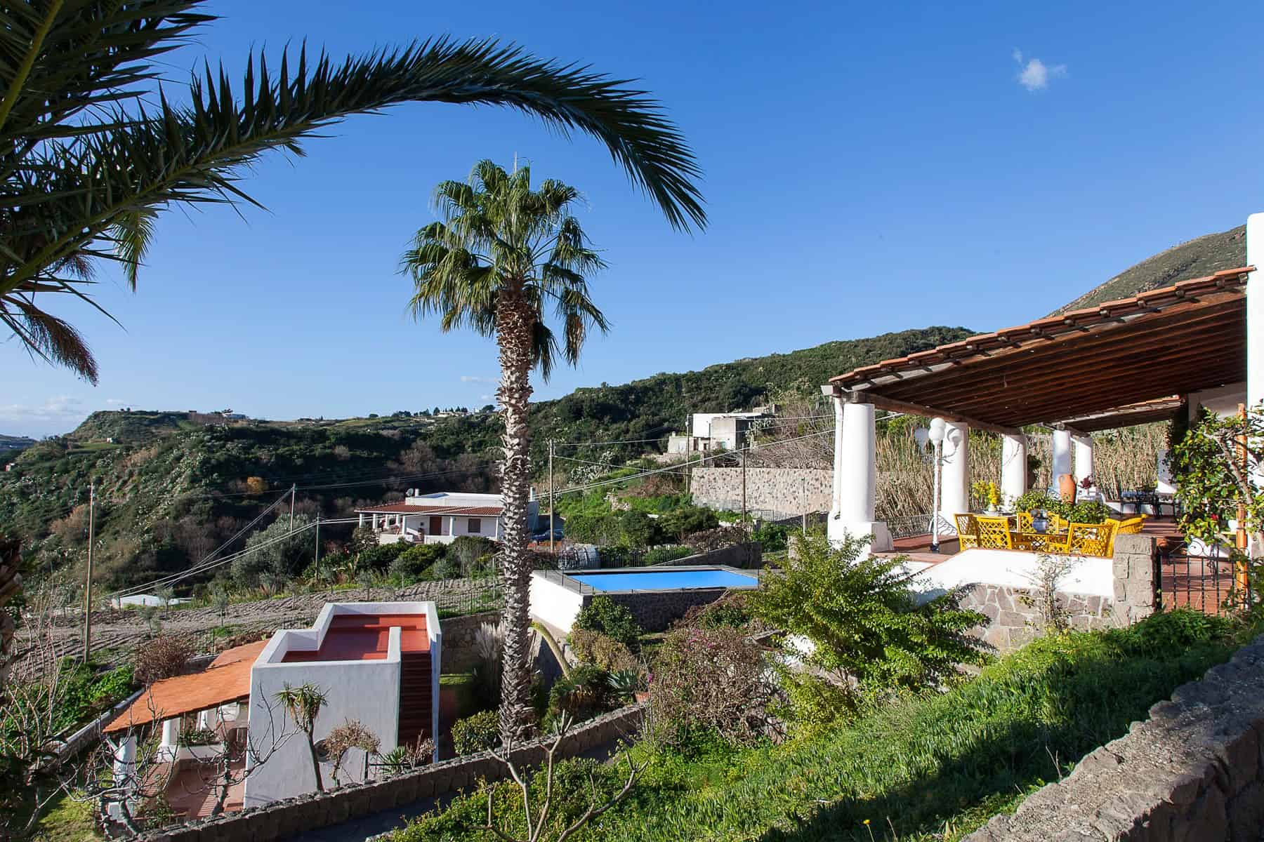 Vista panoramica di una casa con piscina e terrazza all'aperto, circondata da vegetazione e palme, in un paesaggio collinare soleggiato.