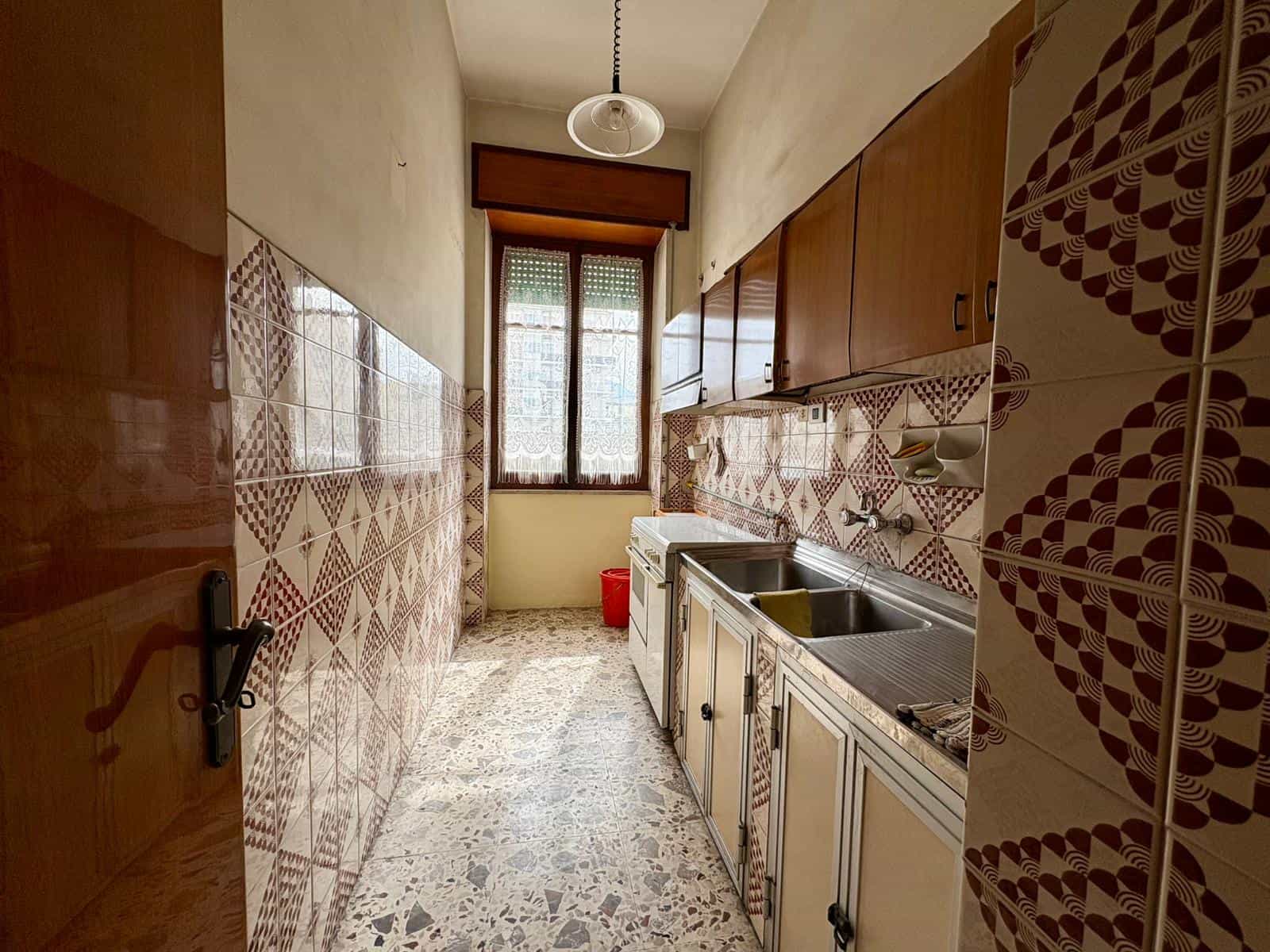 Lavabo e cucina in un appartamento con pareti rivestite di piastrelle decorative e pavimento in marmo, illuminato da una grande finestra che permette l'ingresso di luce naturale.