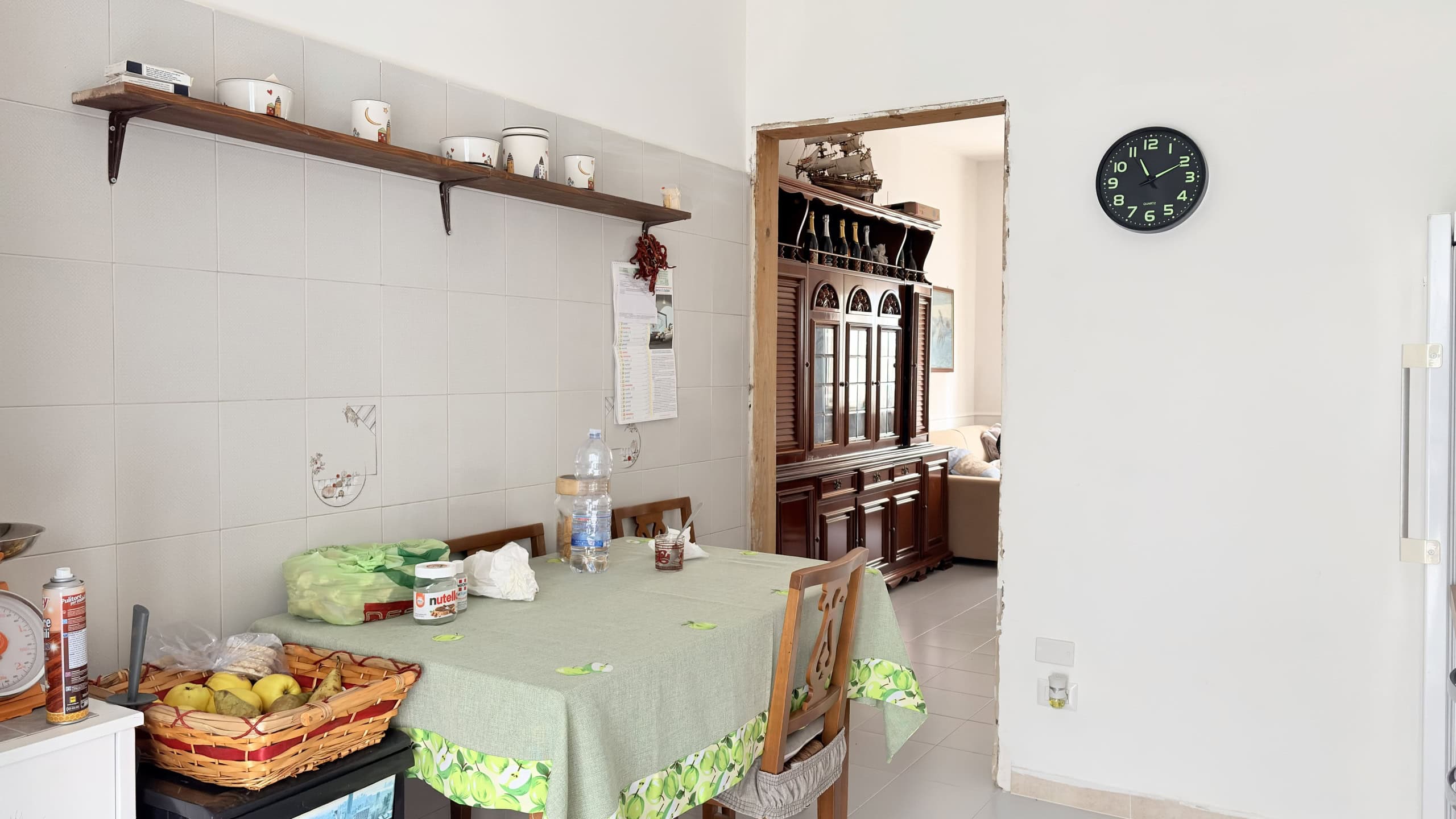 Piatto di frutta fresca e snack su tavolo in cucina luminosa con pareti bianche e scaffale con oggetti decorativi.