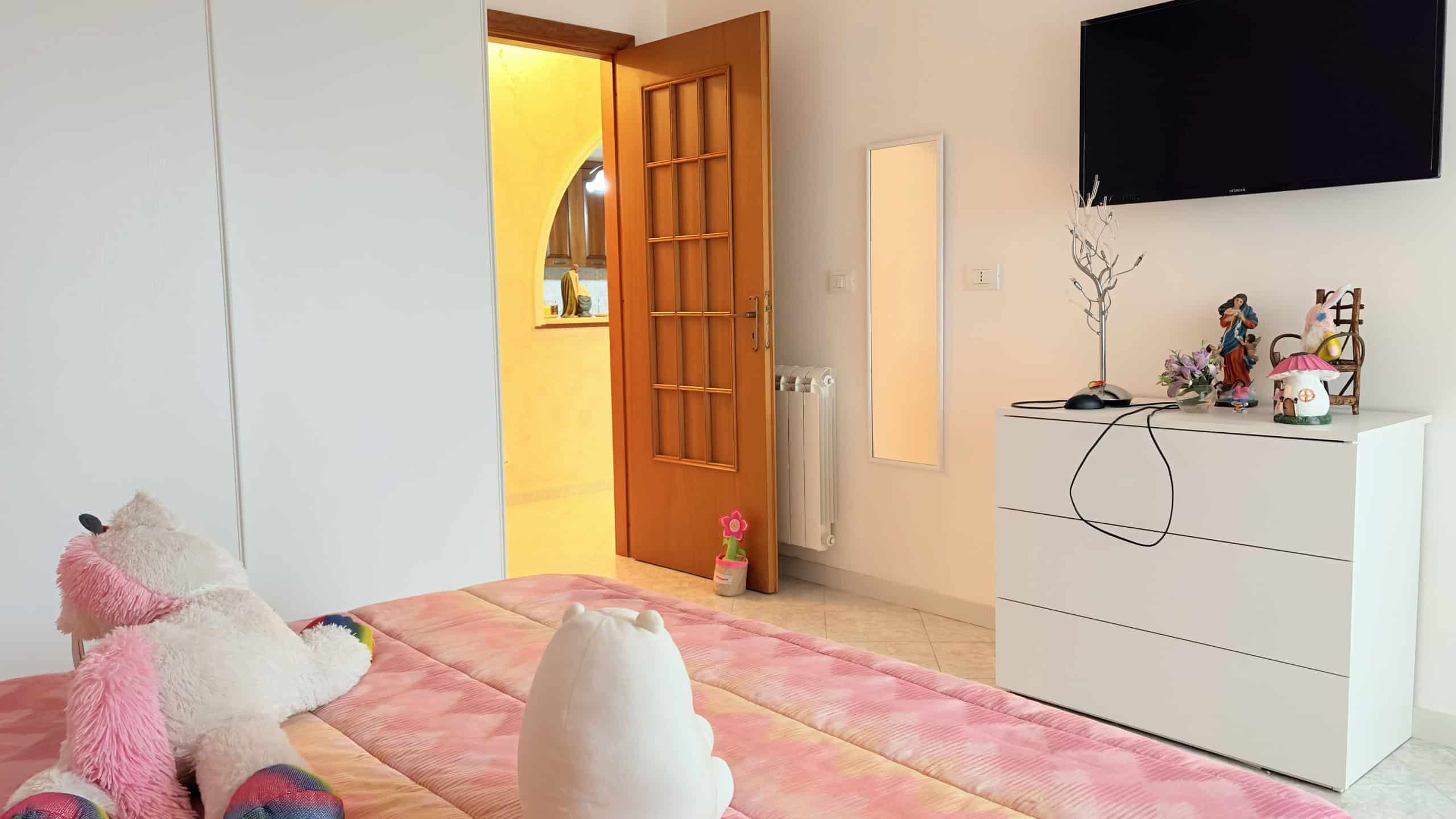 Ampia camera da letto moderna con pareti bianche, letto con copertura rosa e decorazioni morbide, TV a schermo piatto e mobile bianco con oggetti decorativi, ambiente luminoso e accogliente.