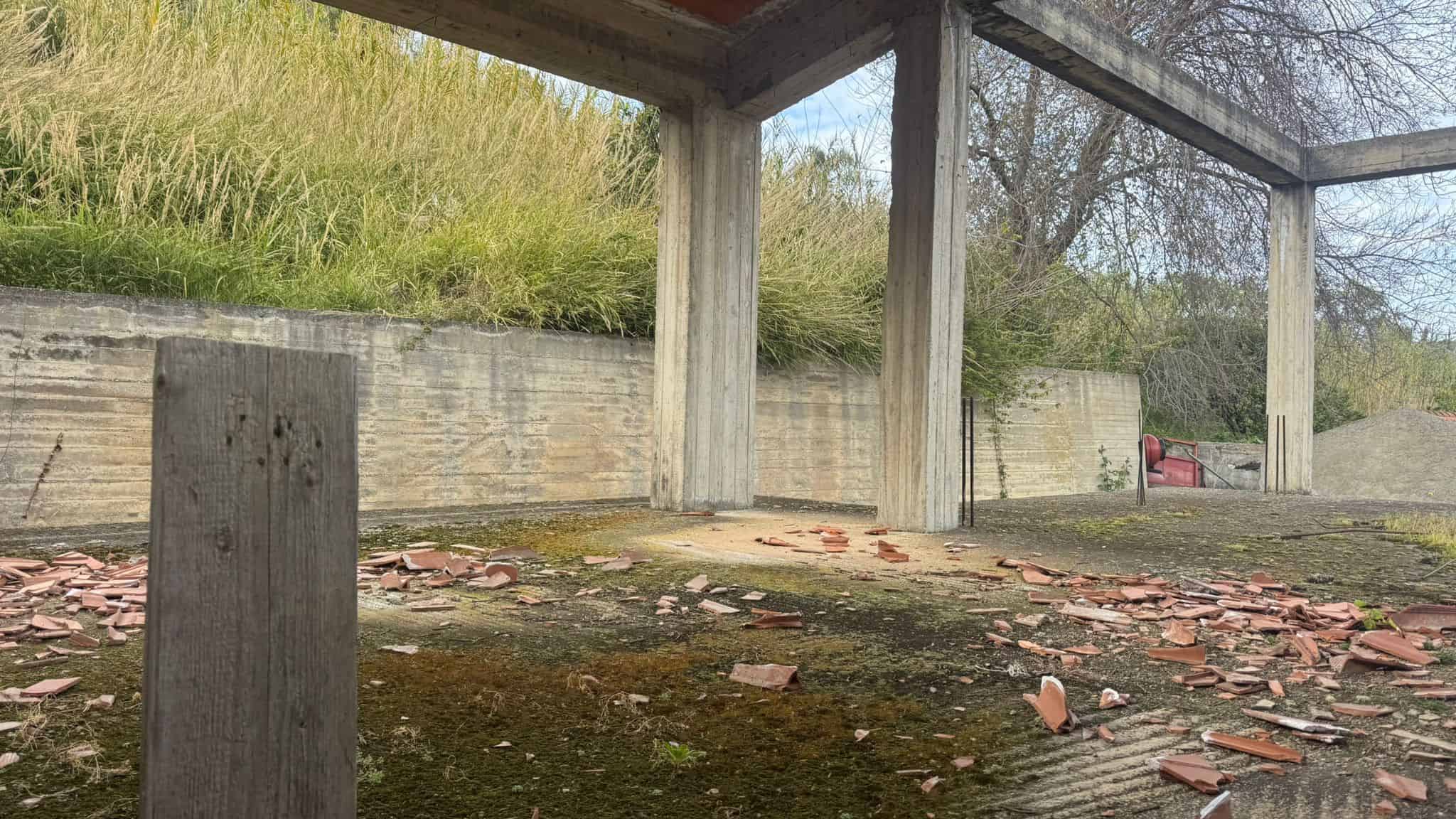 Pavimentazione esterna in pietra con mattoni rotti e struttura in cemento, ambiente all'aperto con vegetazione e pareti di recinzione in legno e cemento.
