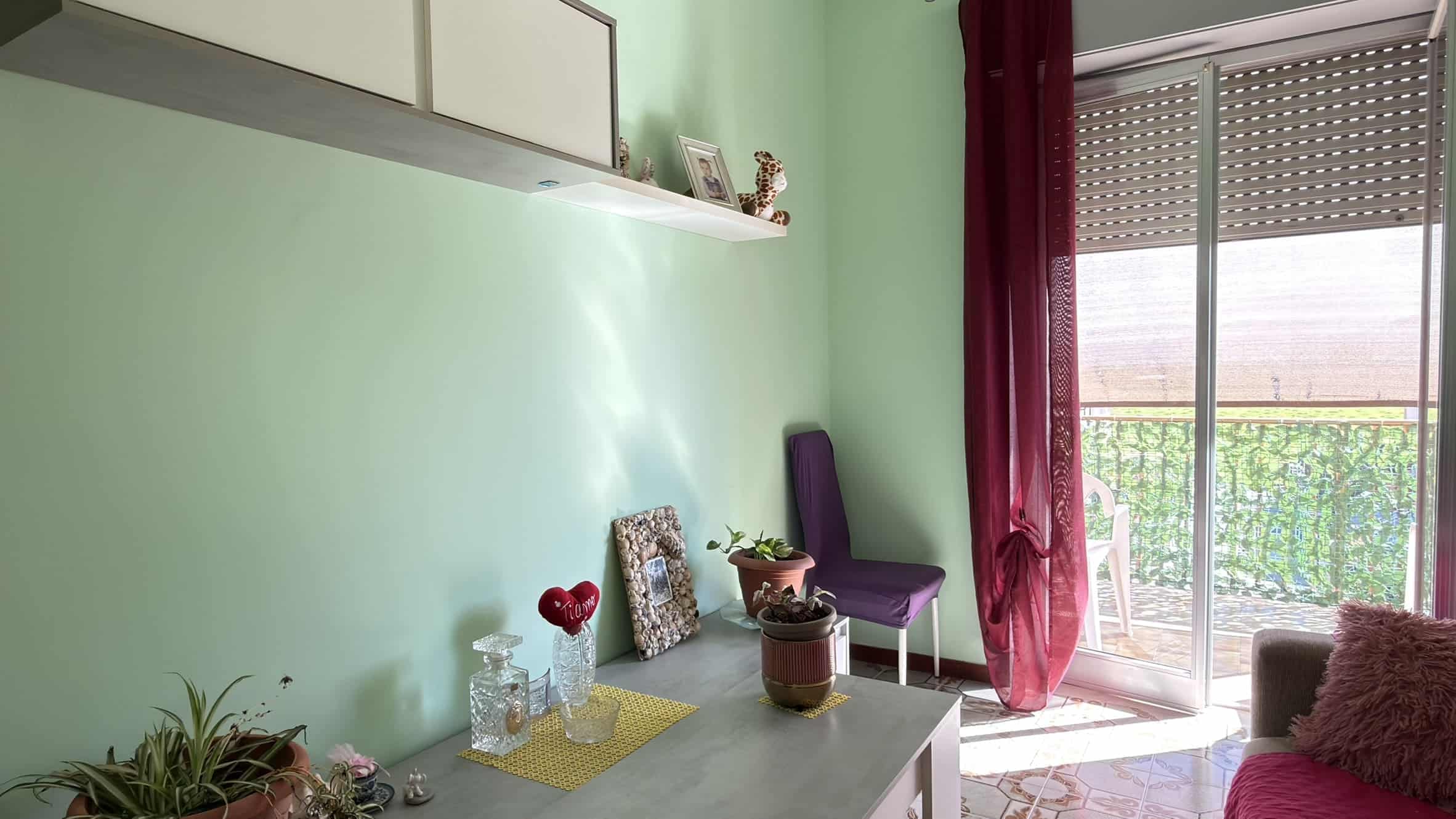 Porte scorrevoli in vetro che conducono a un balcone con vista su un giardino, illuminato dalla luce naturale, in una stanza con pareti verde pastello, decorazioni eleganti e mobili moderni.