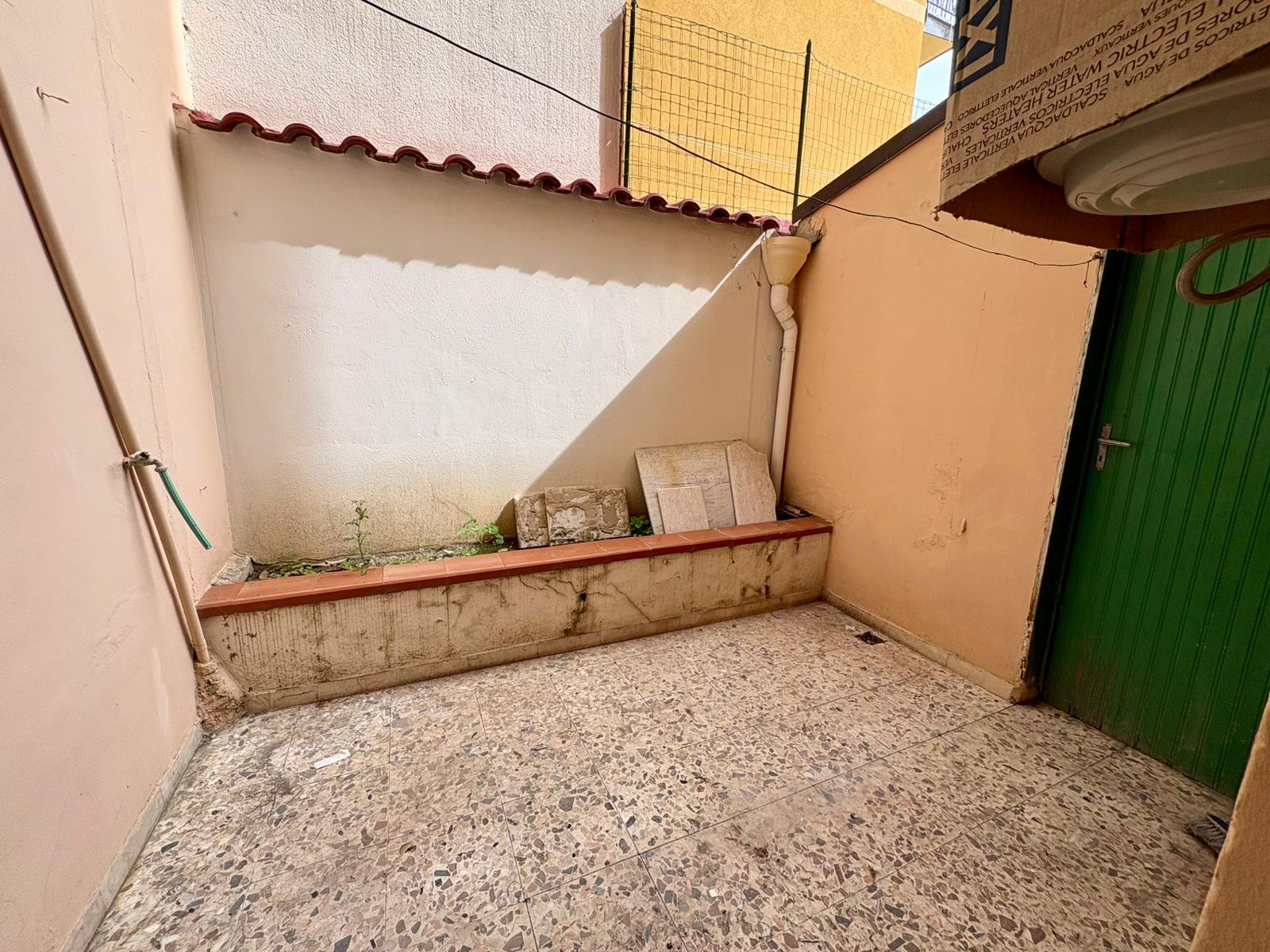 Targhetta di un cortile esterno con pareti rosa, pavimento in pietra e una porta verde, con alcuni mattoni e tubature visibili, in un ambiente residenziale all'aperto.