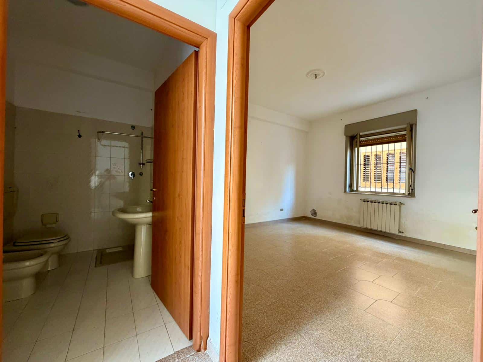 Bagno e soggiorno spazioso in un appartamento in affitto, con pareti bianche e pavimenti in piastrelle, finestra con tende e radiatore, ambiente luminoso e pronto per essere arredato.