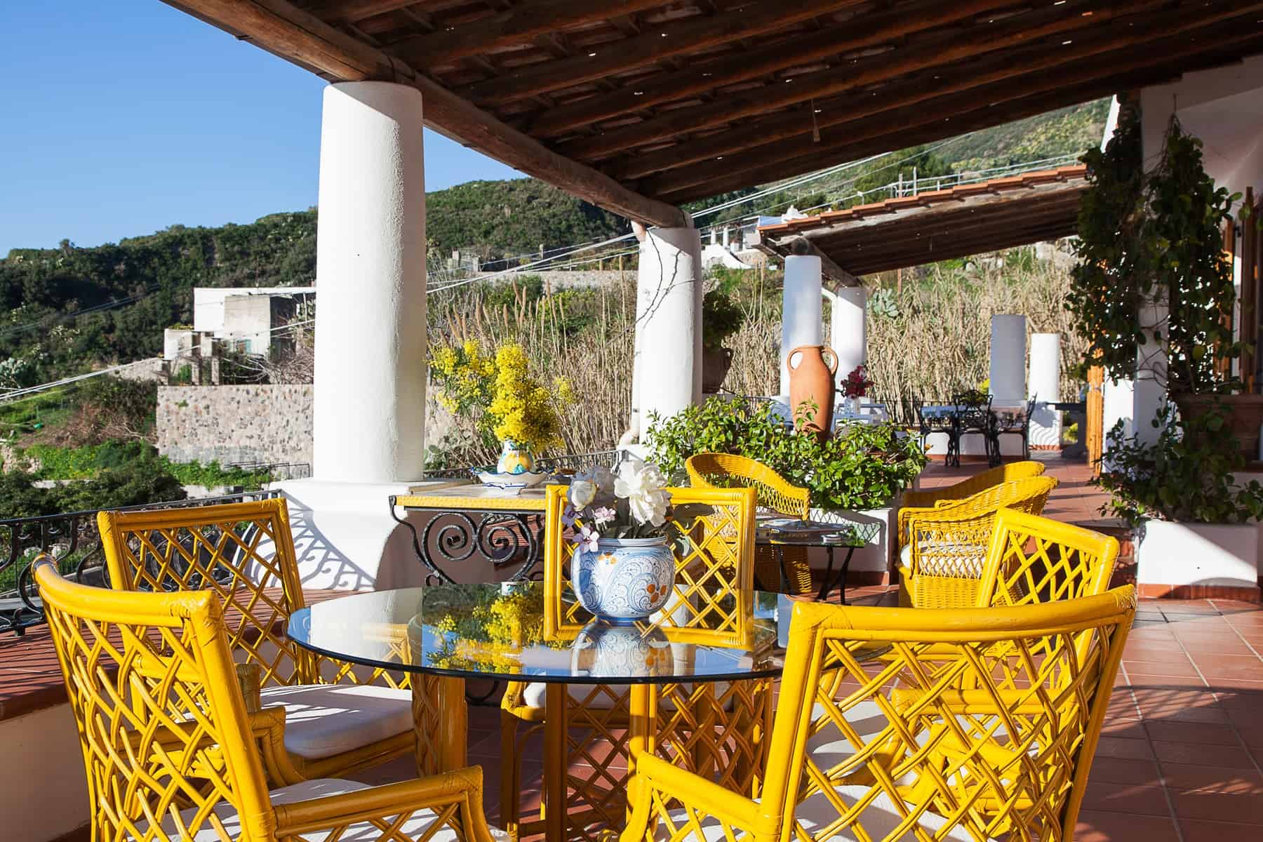Balcone con mobili da esterno in stile mediterraneo, sedie gialle e tavolo in vetro, vista su paesaggio collinare e vegetazione, ambiente luminoso e accogliente.