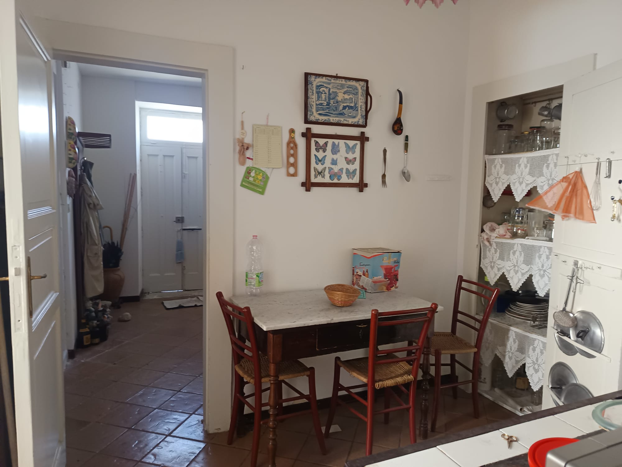 Interno cucina con tavolo in legno, sedie, decorazioni vintage e dispensa con stoviglie, in stile accogliente e tradizionale.
