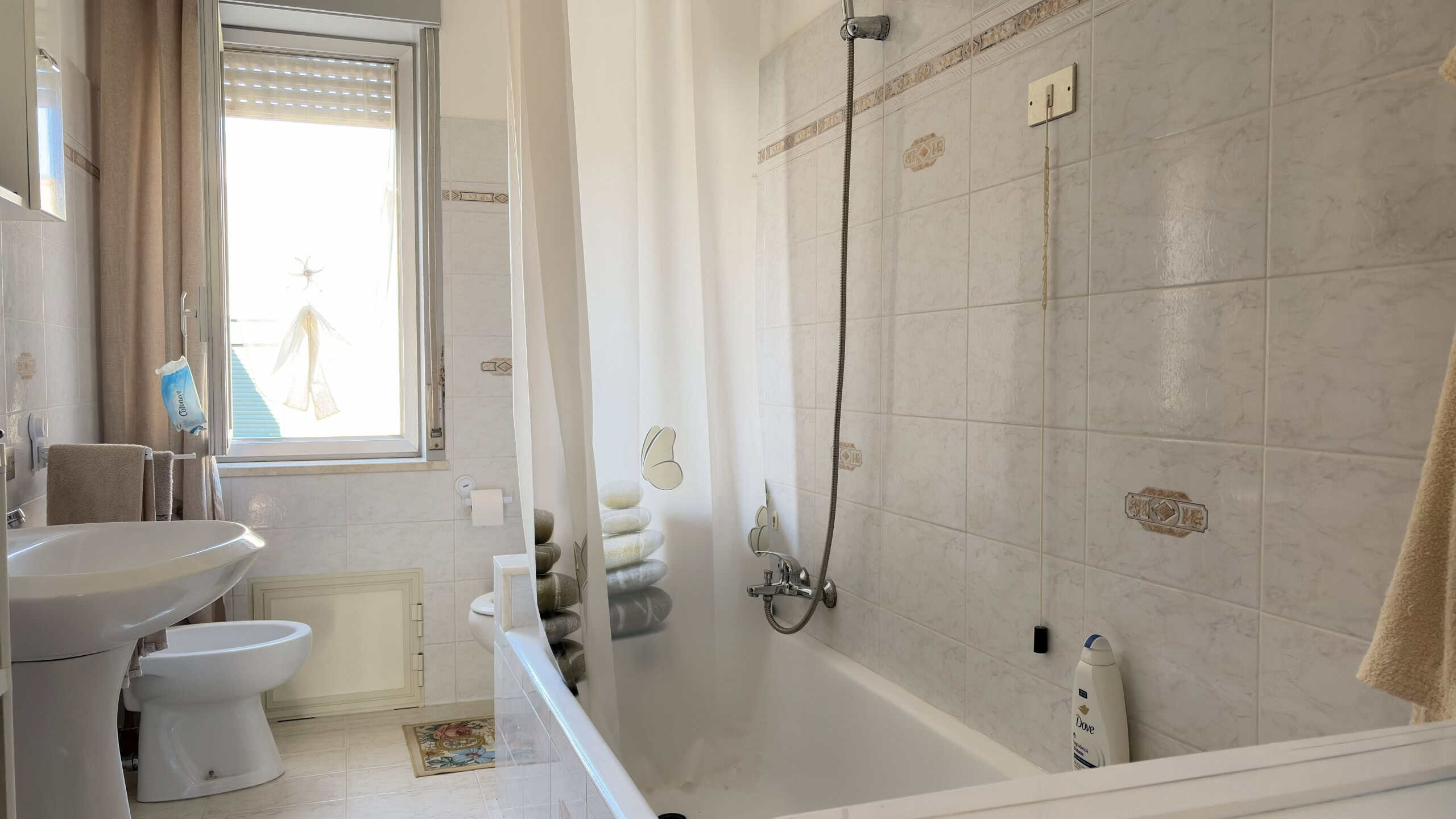 Lavabo, bidet e vasca da bagno in un bagno luminoso con finestra, piastrelle chiare e accessori essenziali, ideale per ambienti di comfort e funzionalità.