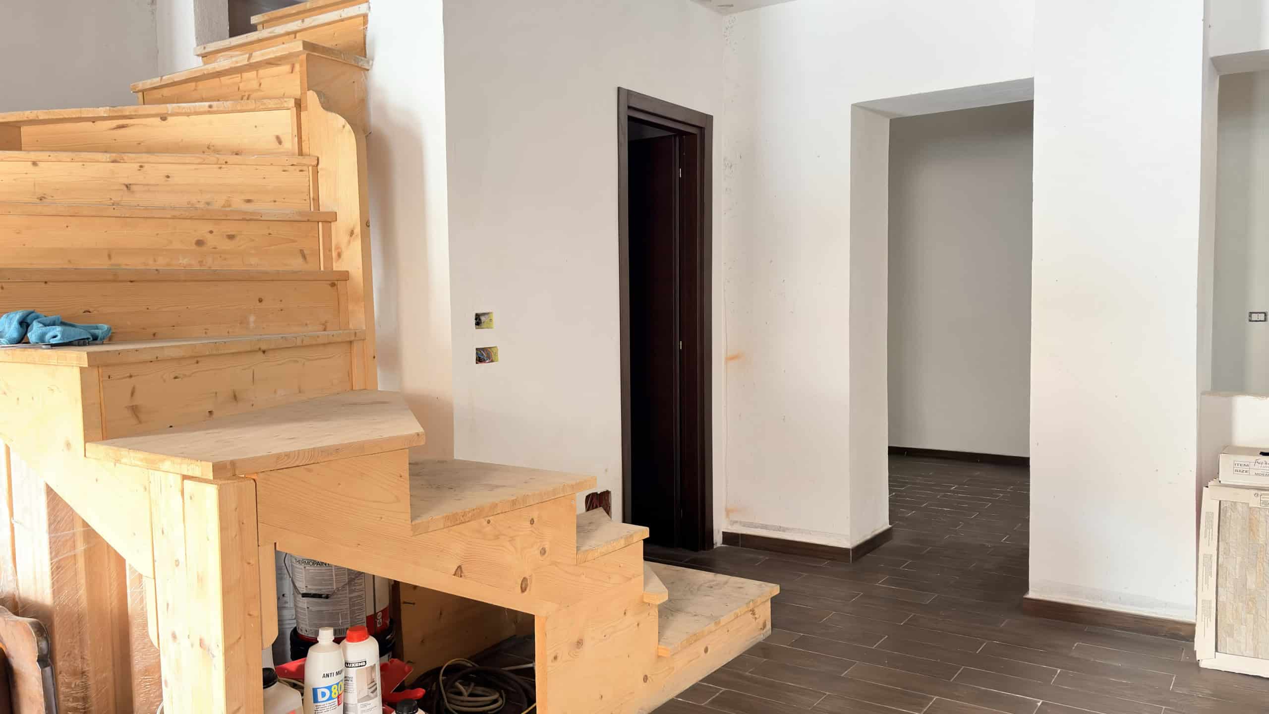 Piantina di una scala in legno moderna all'interno di un'abitazione, con pareti bianche e pavimento in piastrelle scure, pronta per essere installata o ristrutturata.