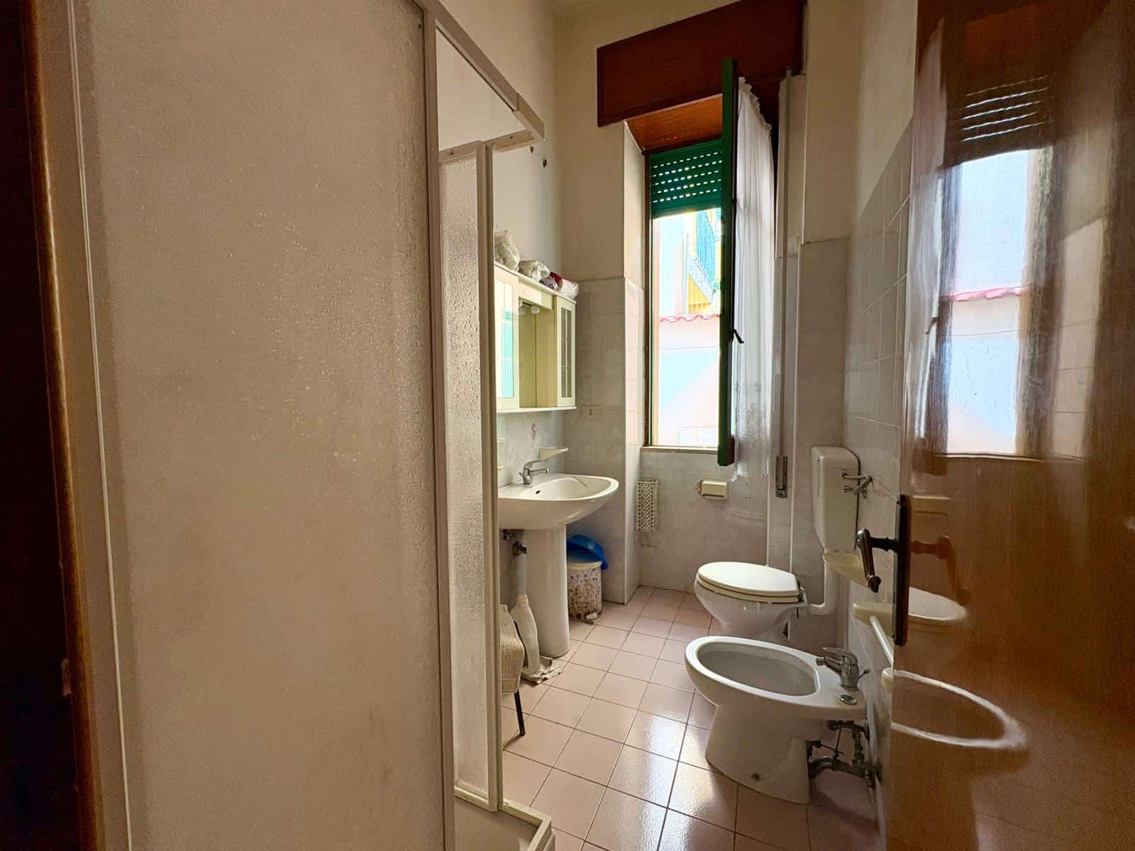 Lavabo e servizi igienici in bagno spazioso con finestra, piastrelle chiare e arredi funzionali, ideale per abitazioni confortevoli e pratiche.
