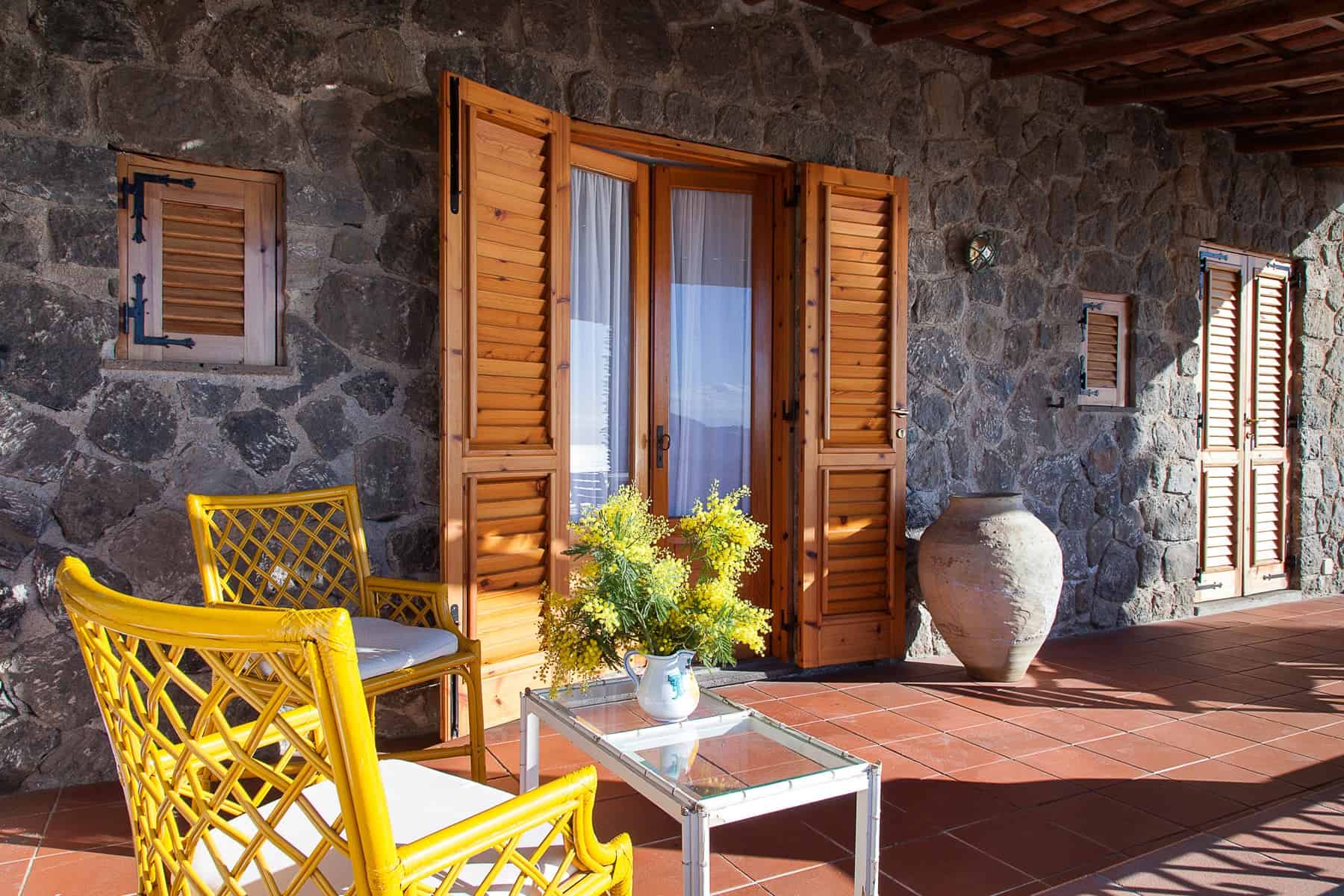 Il testo dell'immagine mostra un'area esterna di una casa in pietra con porte e finestre in legno, arredamento da esterno con sedie gialle e un tavolino, e piante decorative.