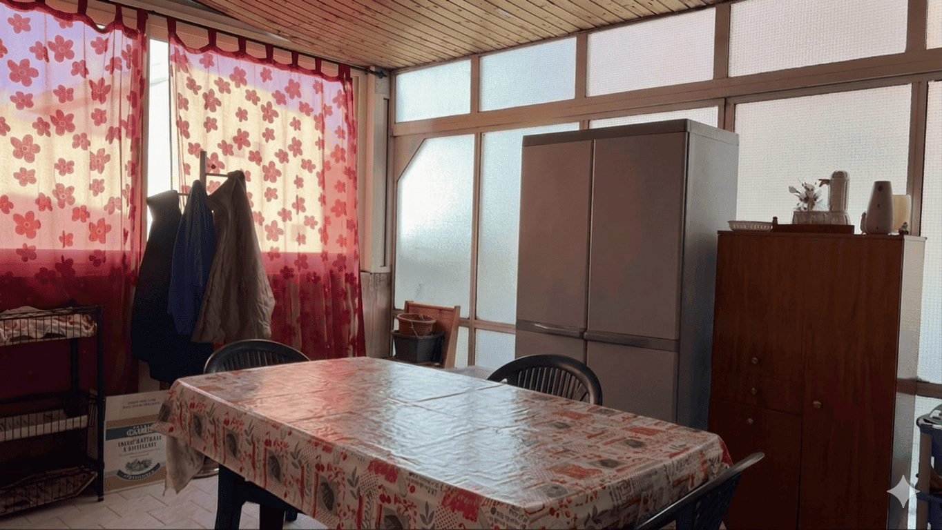 Interno di una cucina con tavolo coperto da tovaglia floreale, frigorifero e mobili in legno, illuminata dalla luce naturale attraverso grandi finestre con tende rosse a fiori.