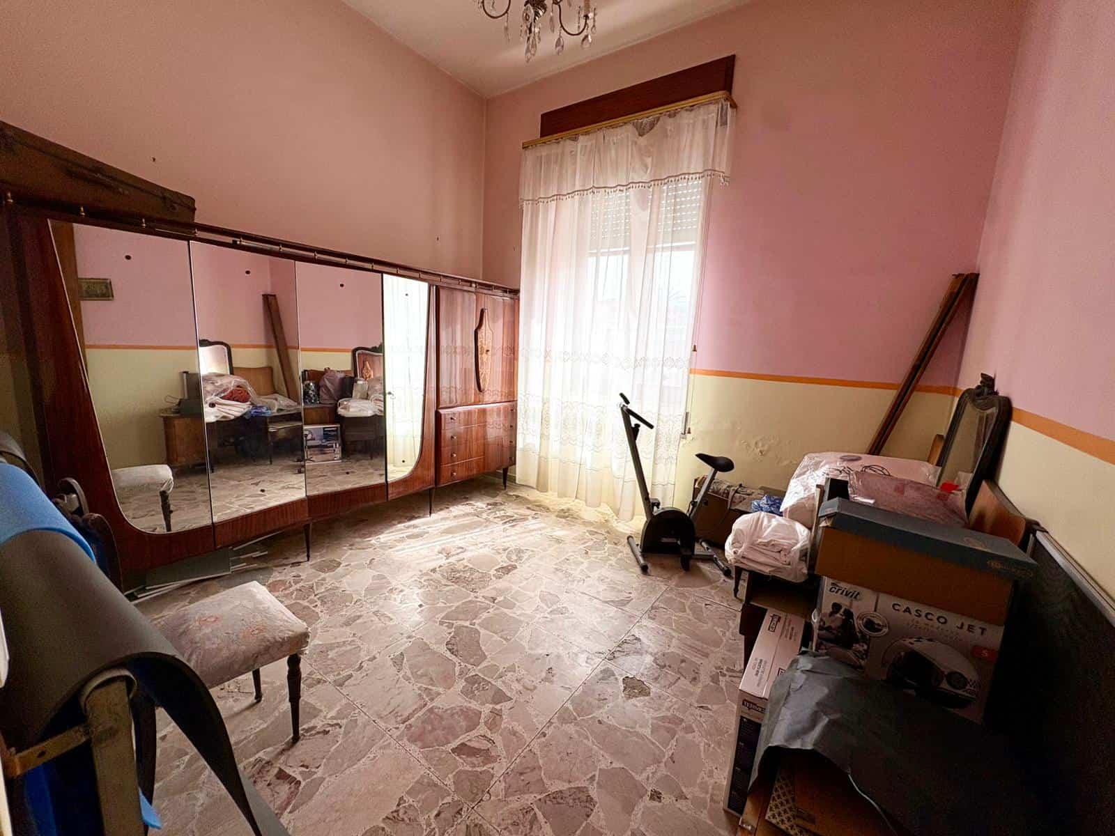 Camera da letto vuota con armadio a specchio, sedia, e finestra con tende leggere, pavimento in marmo, ambiente luminoso e semplice.